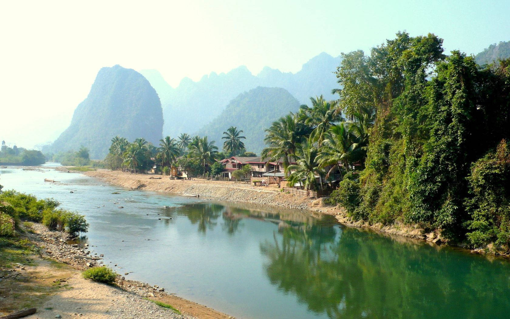 Laos 桌布 壁紙 — 一條寧靜的河流蜿蜒流過茂密的綠植和棕櫚樹，遠處的喀斯特山脈在薄霧中聳立。