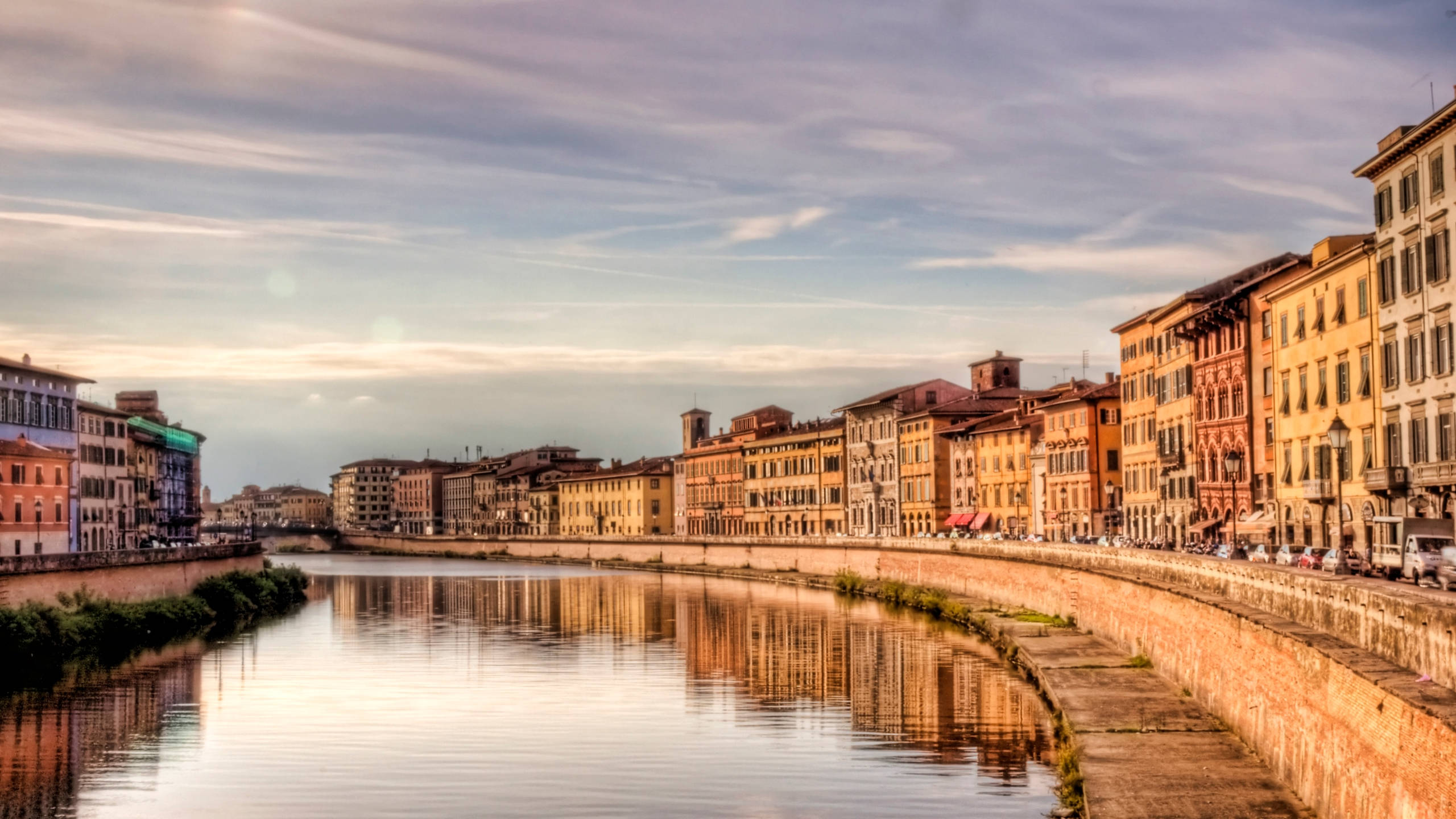 Tuscany 桌布 壁紙 — 一條寧靜的河流蜿蜒穿過一座歷史悠久的歐洲城市，色彩繽紛的建築物倒映在平靜的水面上，天空柔和。