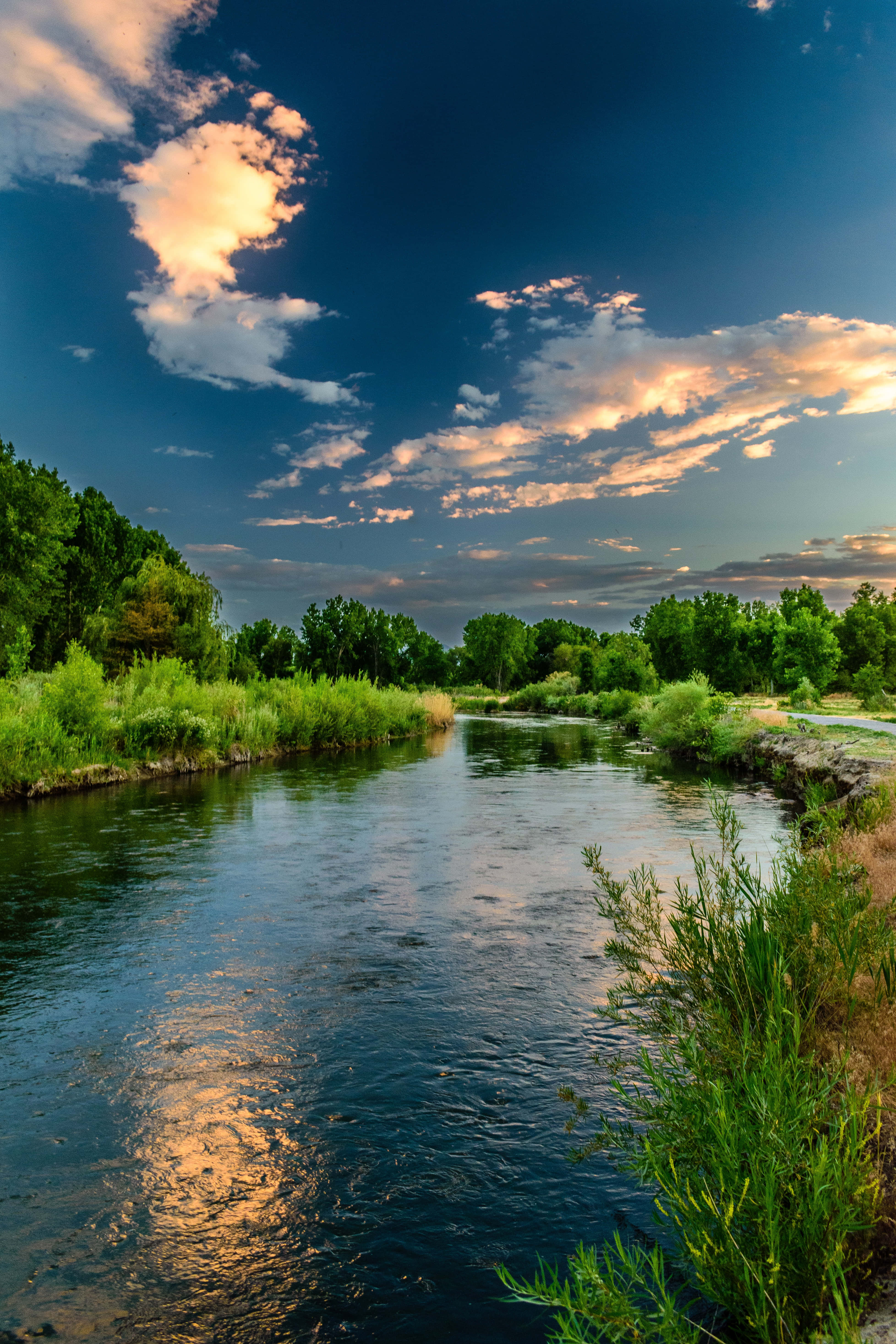 river 桌布 壁紙 — 一條寧靜的河流蜿蜒穿過茂密的綠色河岸，在金色雲彩的戲劇性日落天空下。