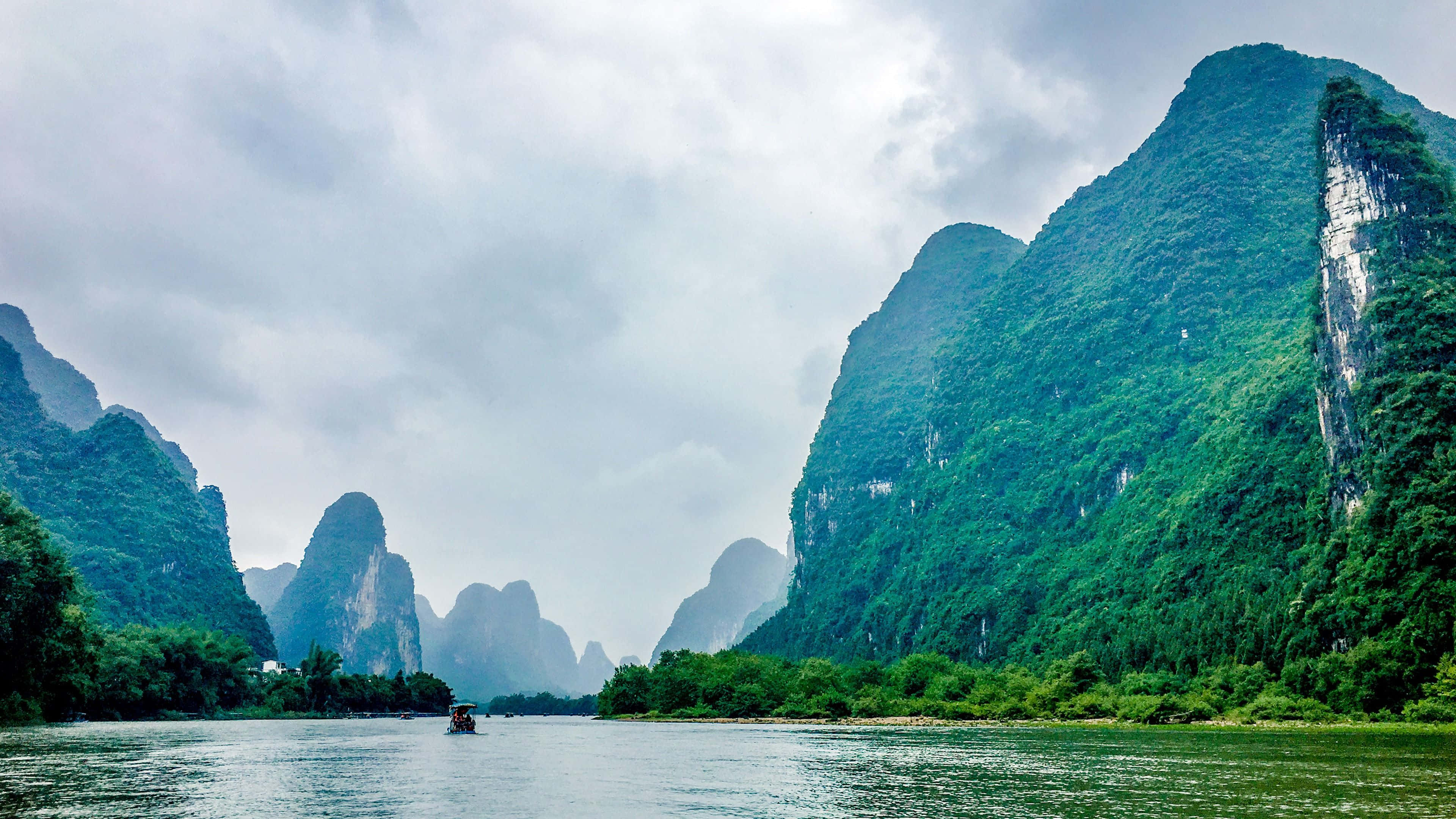 Beautiful Travel 桌布 壁紙 — 一條寧靜的河流蜿蜒穿過鬱鬱蔥蔥的綠色喀斯特山脈，天空多雲，遠處有一艘小船漂流。
