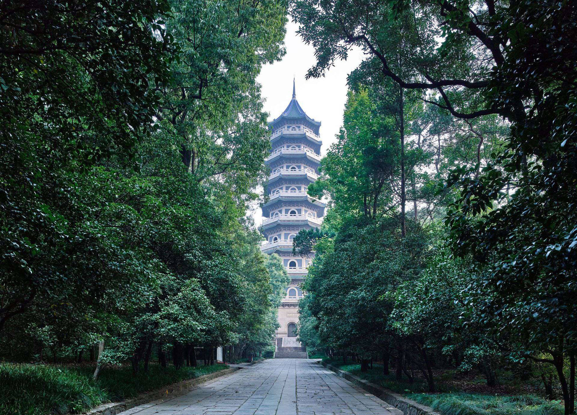 Nanjing 桌布 壁紙 — 一條寧靜的石徑蜿蜒穿過茂密的綠樹，通往一座傳統的多層寶塔，天空多雲。