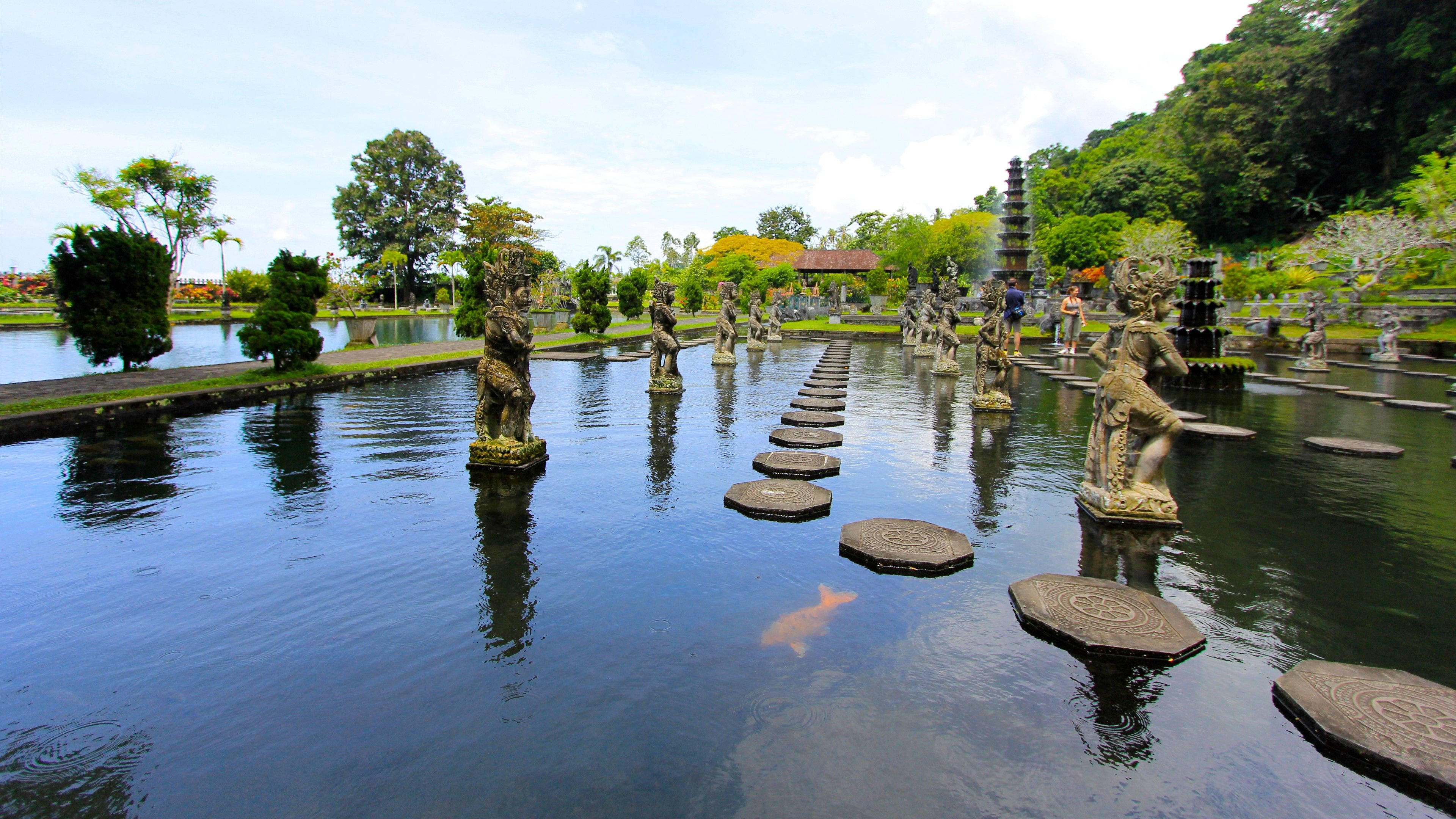 Jakarta 桌布 壁紙 — 一條寧靜的石板小徑穿過平靜的池塘，池塘邊點綴著華麗的雕像，上方是明亮的天空。