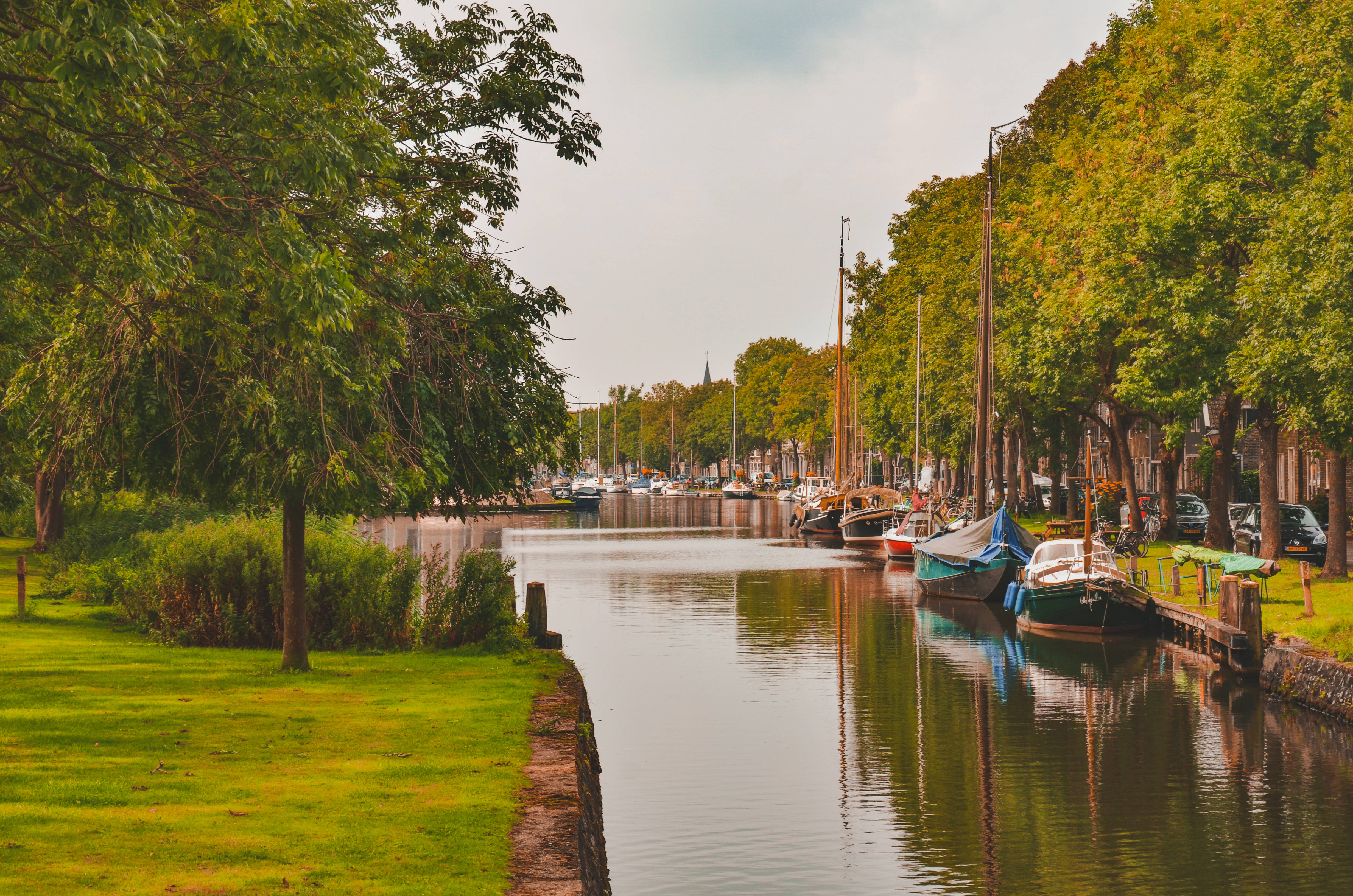 Netherlands 桌布 壁紙 — 一條寧靜的運河，兩旁綠樹成蔭，停泊的帆船在柔和的多雲天空下。