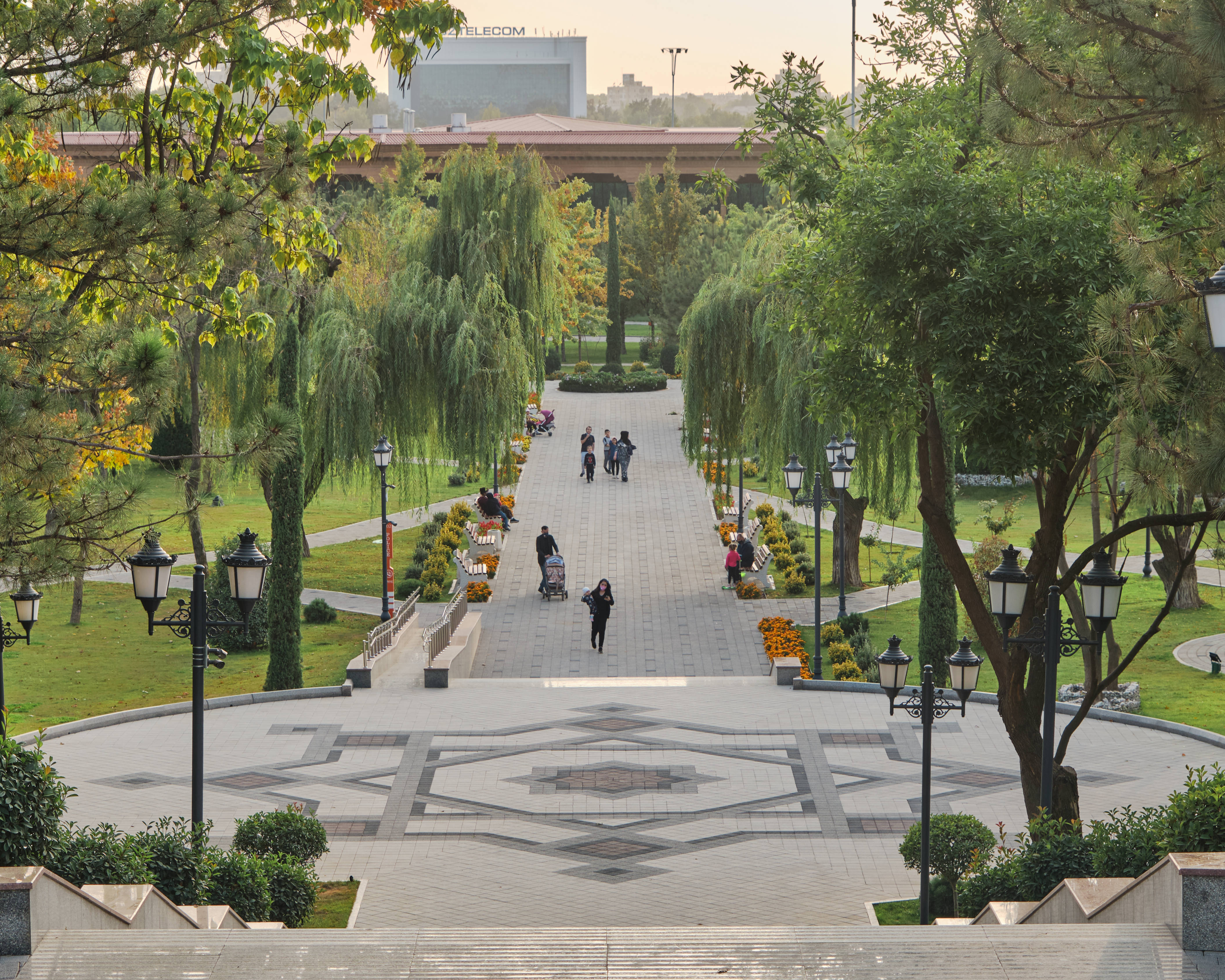 Uzbekistan 桌布 壁紙 — 一條寬闊的公園小徑，兩旁樹木成蔭，人們在秋色葉子下漫步或坐在長椅上。
