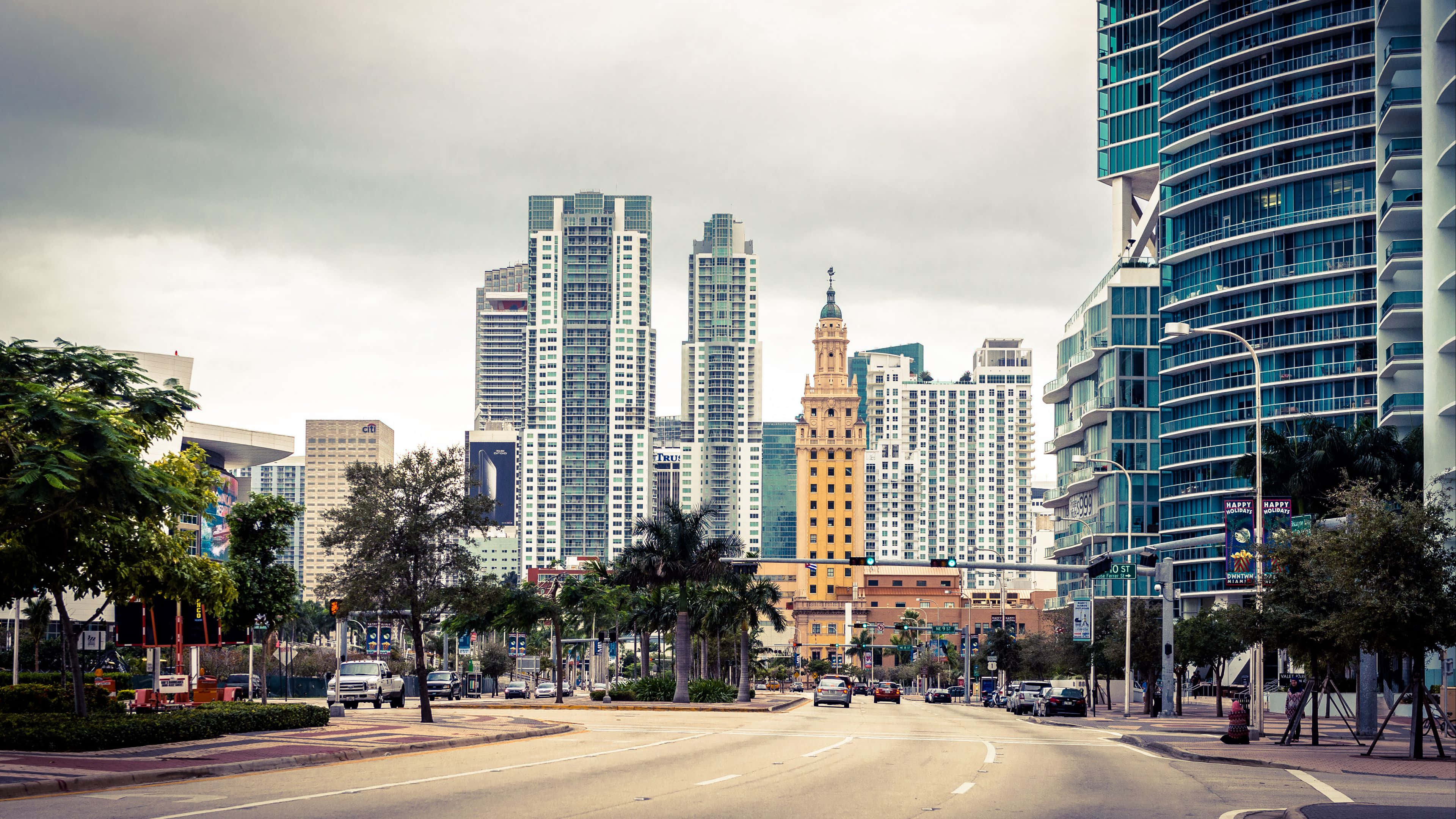 Miami 4K 桌布 壁紙 — 一條寬闊的城市街道兩旁種滿棕櫚樹，通向由現代玻璃塔樓和一座歷史悠久的橙色鐘樓組成的天際線，天空多雲。