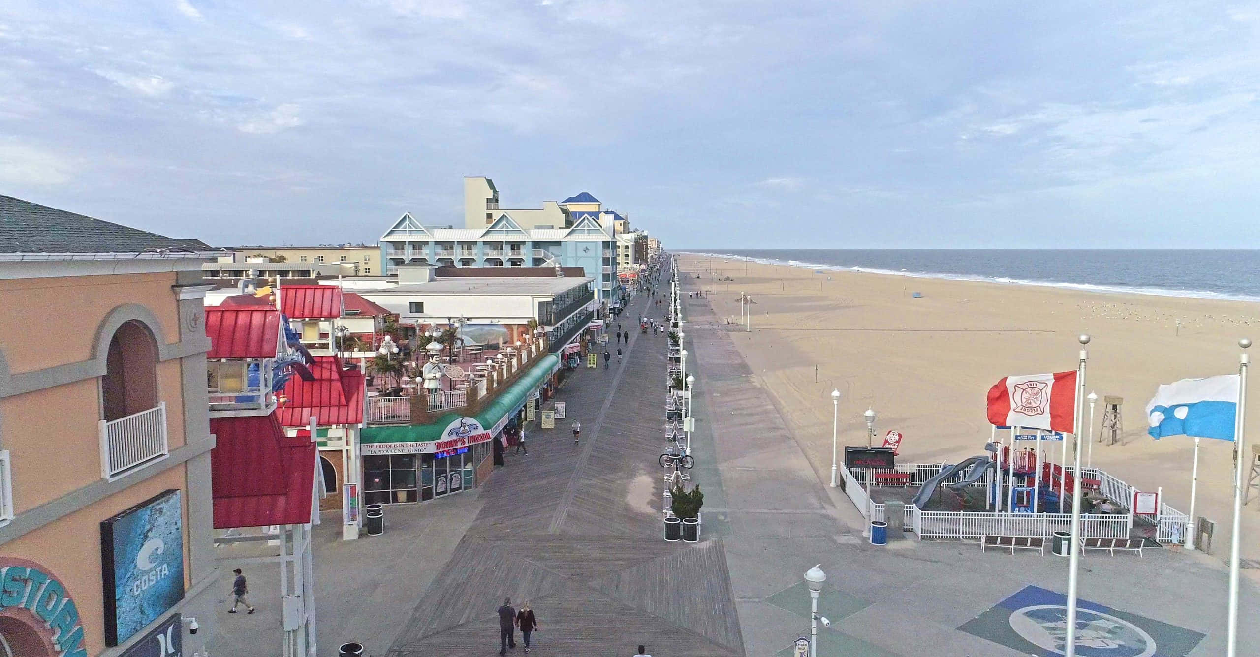 Boardwalk 桌布 壁紙 — 一條寬闊的木板路沿著沙灘延伸，有海浪和色彩繽紛的建築物，天空多雲。