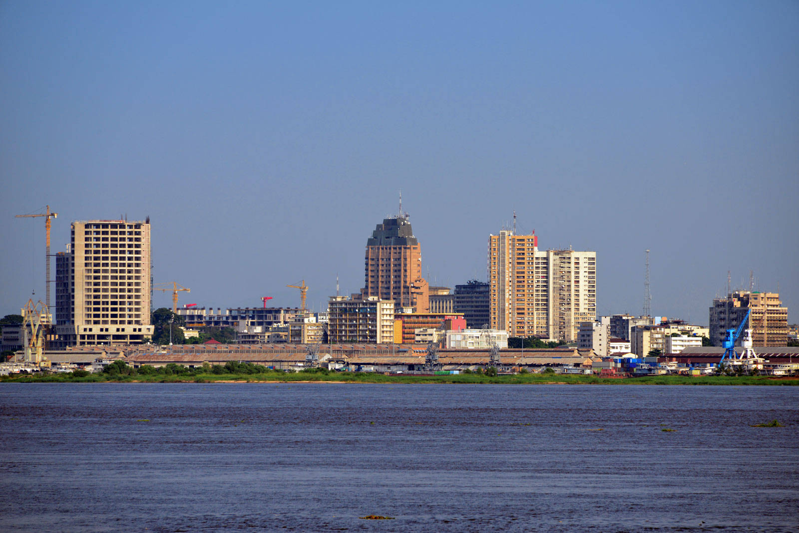 Kinshasa 桌布 壁紙 — 一條寬闊的河流在晴朗的藍天下，延伸於高樓大廈和建築起重機的城市天際線前。