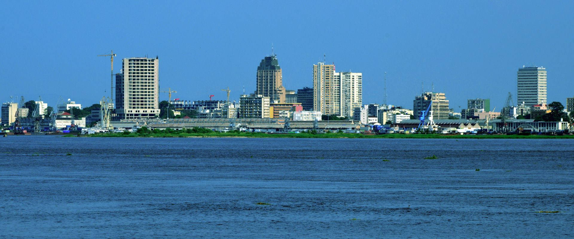 Kinshasa 桌布 壁紙 — 一條寬闊的河流在晴朗的藍天下延伸至現代城市天際線前，建築起重機清晰可見。