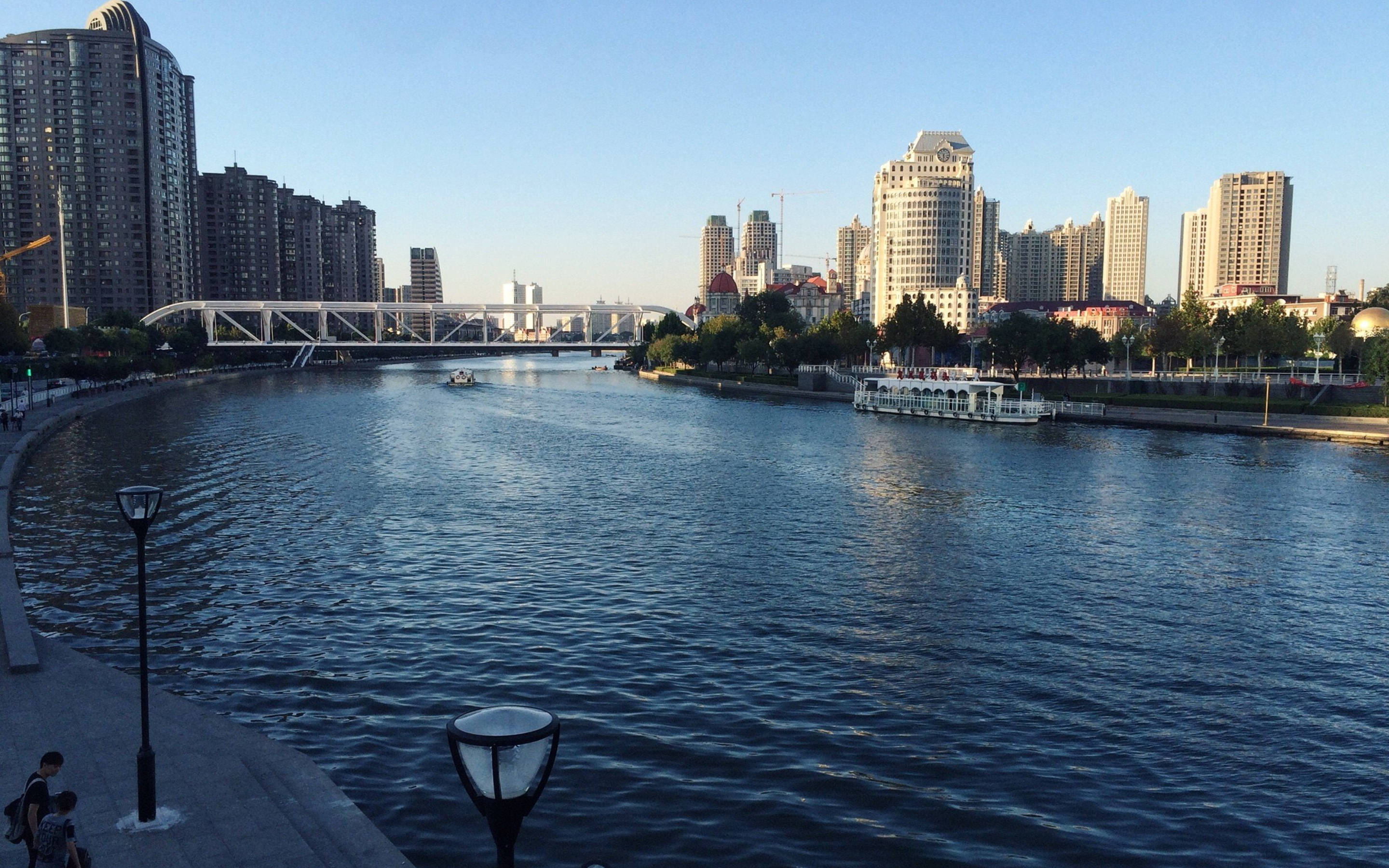Tianjin 桌布 壁紙 — 一條寬闊的河流流經一座現代城市，兩旁高樓大廈林立，一座白色橋樑橫跨其上，天空湛藍清澈。