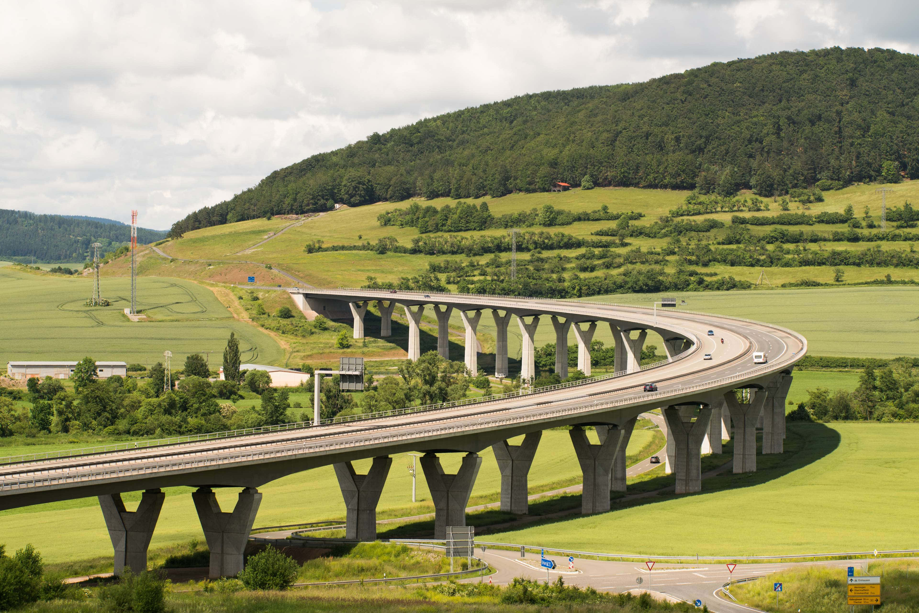 Infrastructure 桌布 壁紙 — 一條寬闊的混凝土高速公路橋在陰天之下優雅地彎曲越過鬱鬱蔥蔥的綠色山丘。