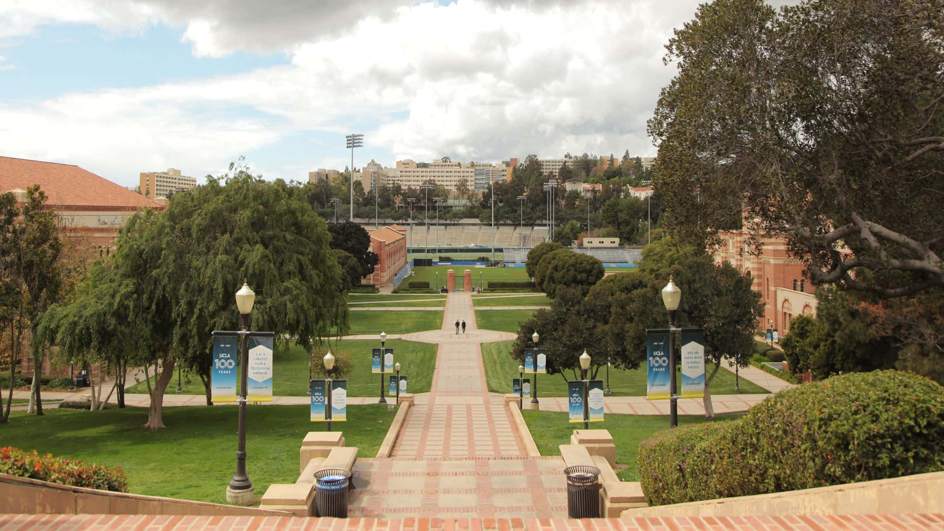 Ucla Campus 桌布 壁紙 — 一條寬闊的石徑穿過綠意盎然的校園，通往一個部分多雲天空下的體育場。