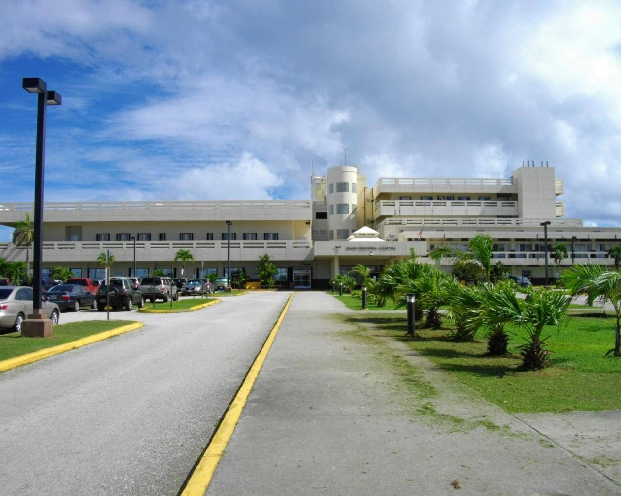 Guam 桌布 壁紙 — 一條寬闊的車道通向一座大型米色醫院建築，天空部分多雲，有棕櫚樹和停放的汽車。