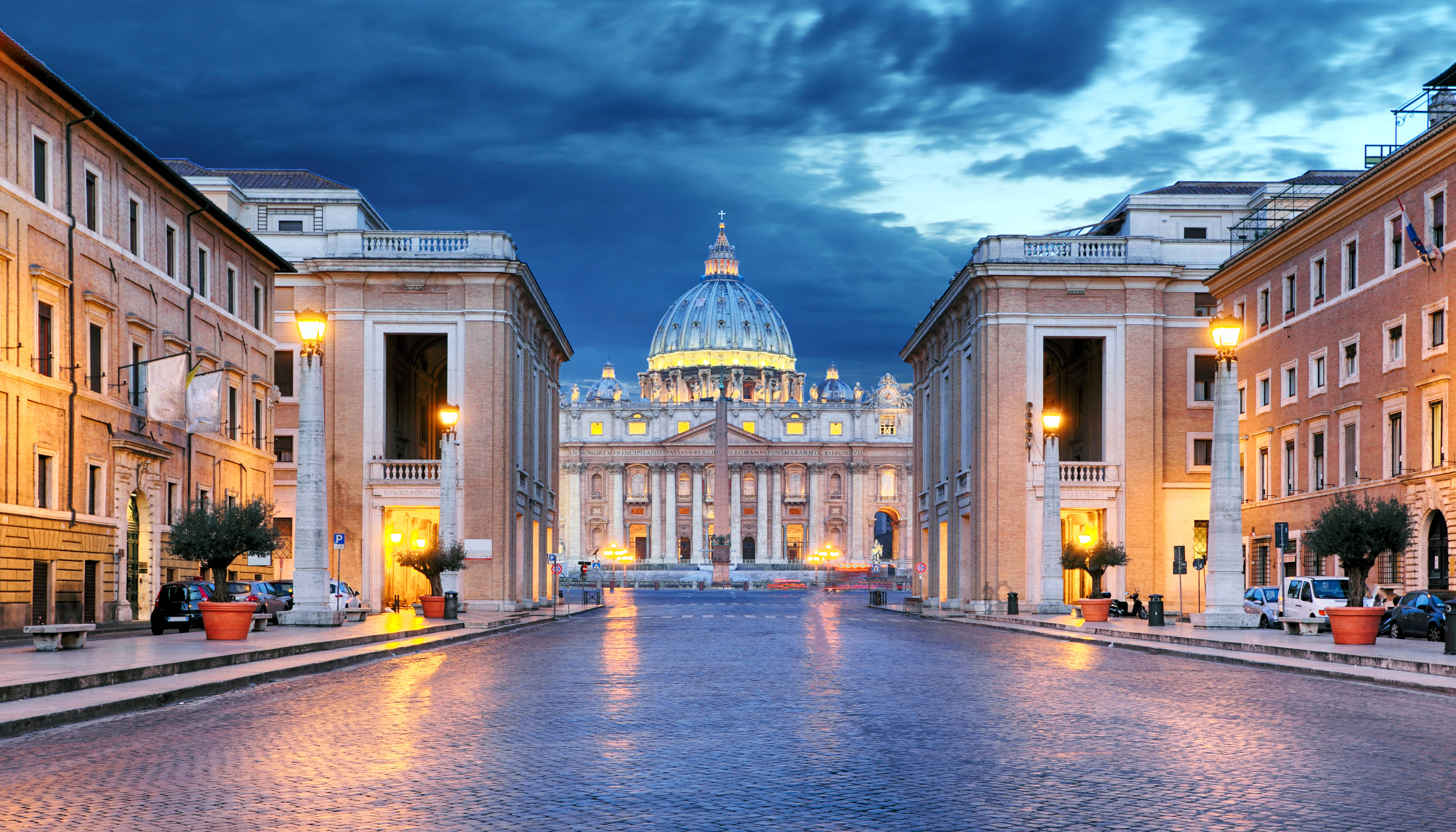 Vatican 桌布 壁紙 — 一條寬闊的鵝卵石大道通往在戲劇性暮色天空下被照亮的聖彼得大教堂圓頂。