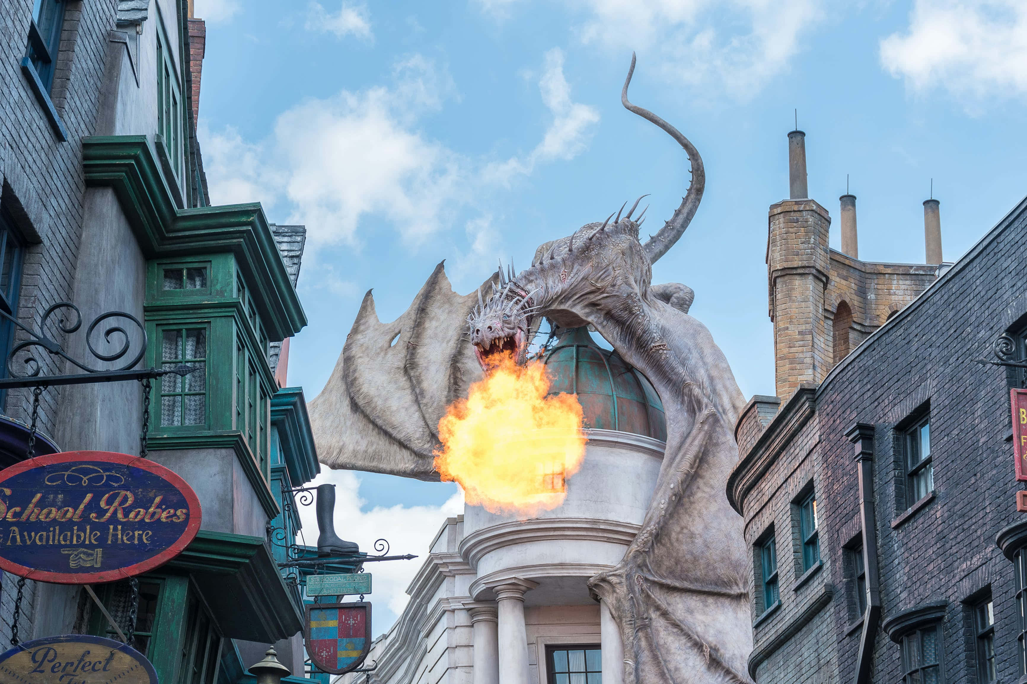 Wizarding World 桌布 壁紙 — 一條巨大的龍在晴朗的藍天下，向一條鋪滿鵝卵石、兩旁有巫師商店的街道噴火。