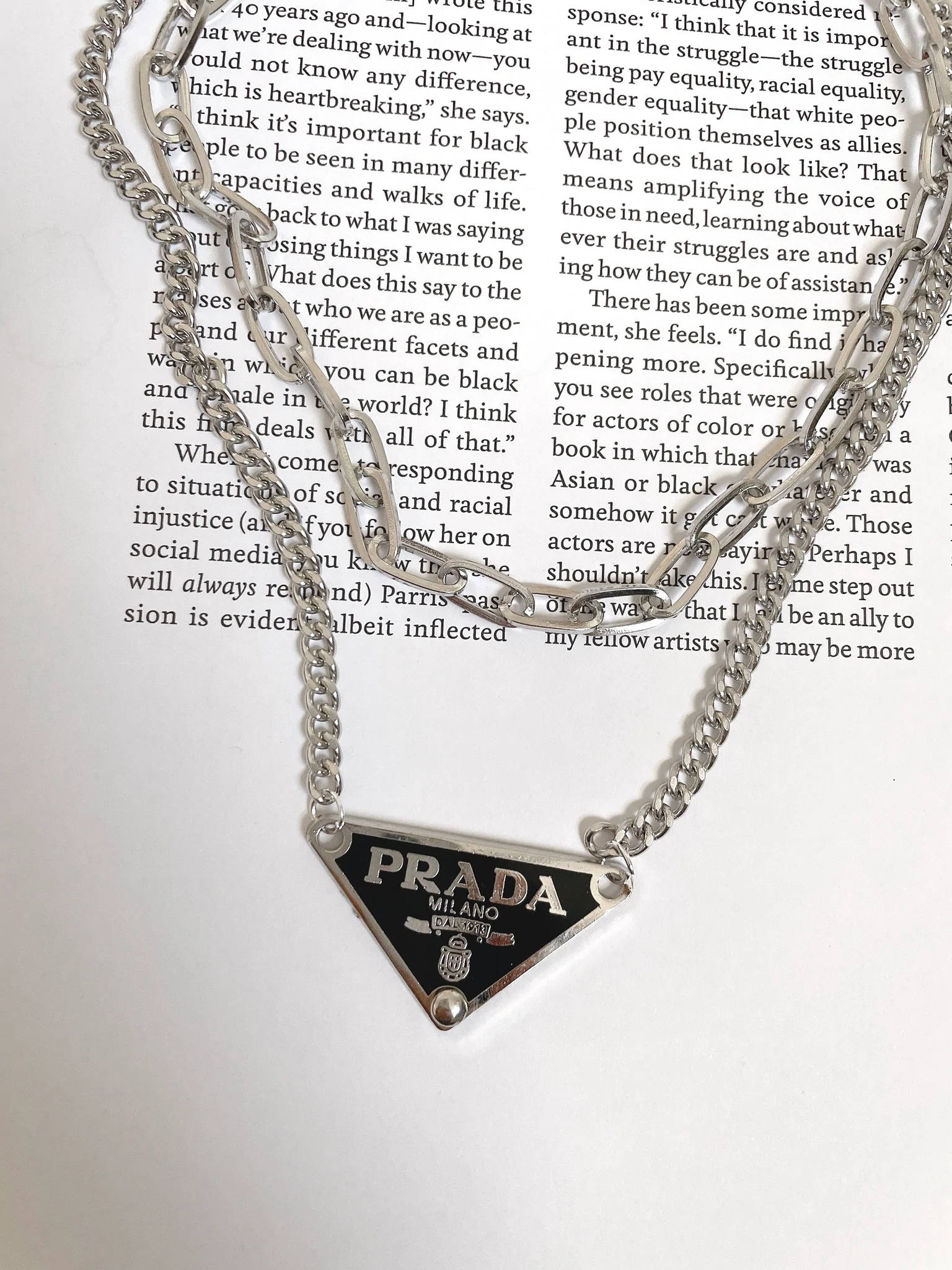 Prada 桌布 壁紙 — 一條帶有三角形標誌標籤的銀色 Prada 鏈條項鍊，放在印有文字的打開書頁上。