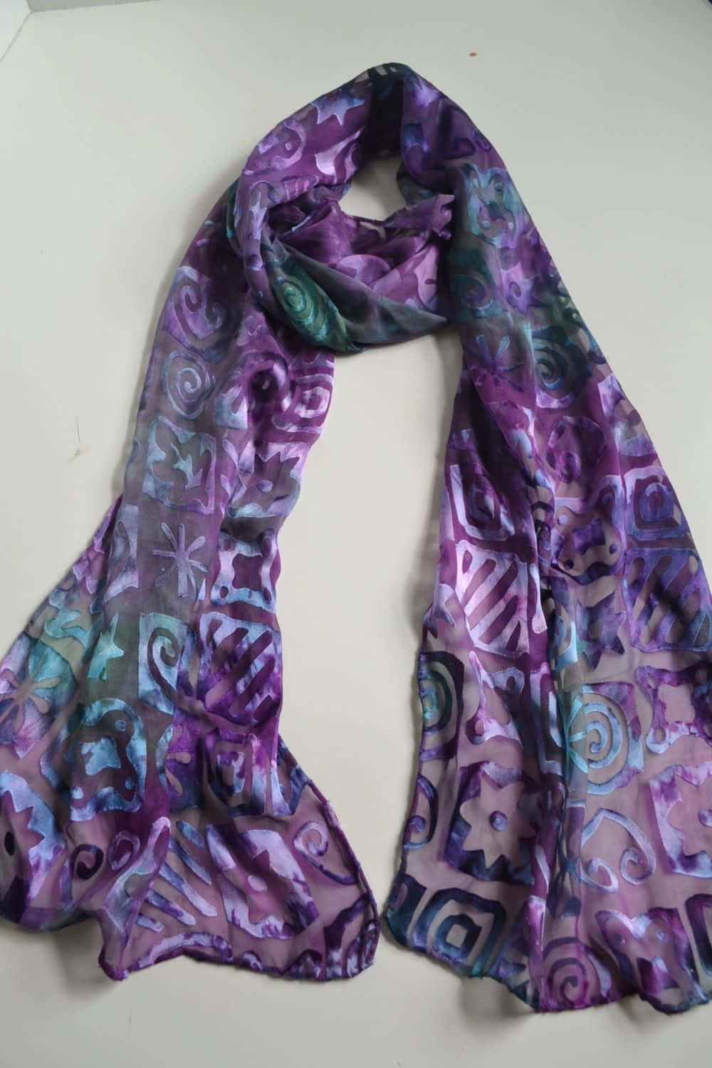 Purple Scarf 桌布 壁紙 — 一條帶有抽象漩渦和幾何圖案的紫色和藍綠色流動圖案圍巾，披在純白色表面上。
