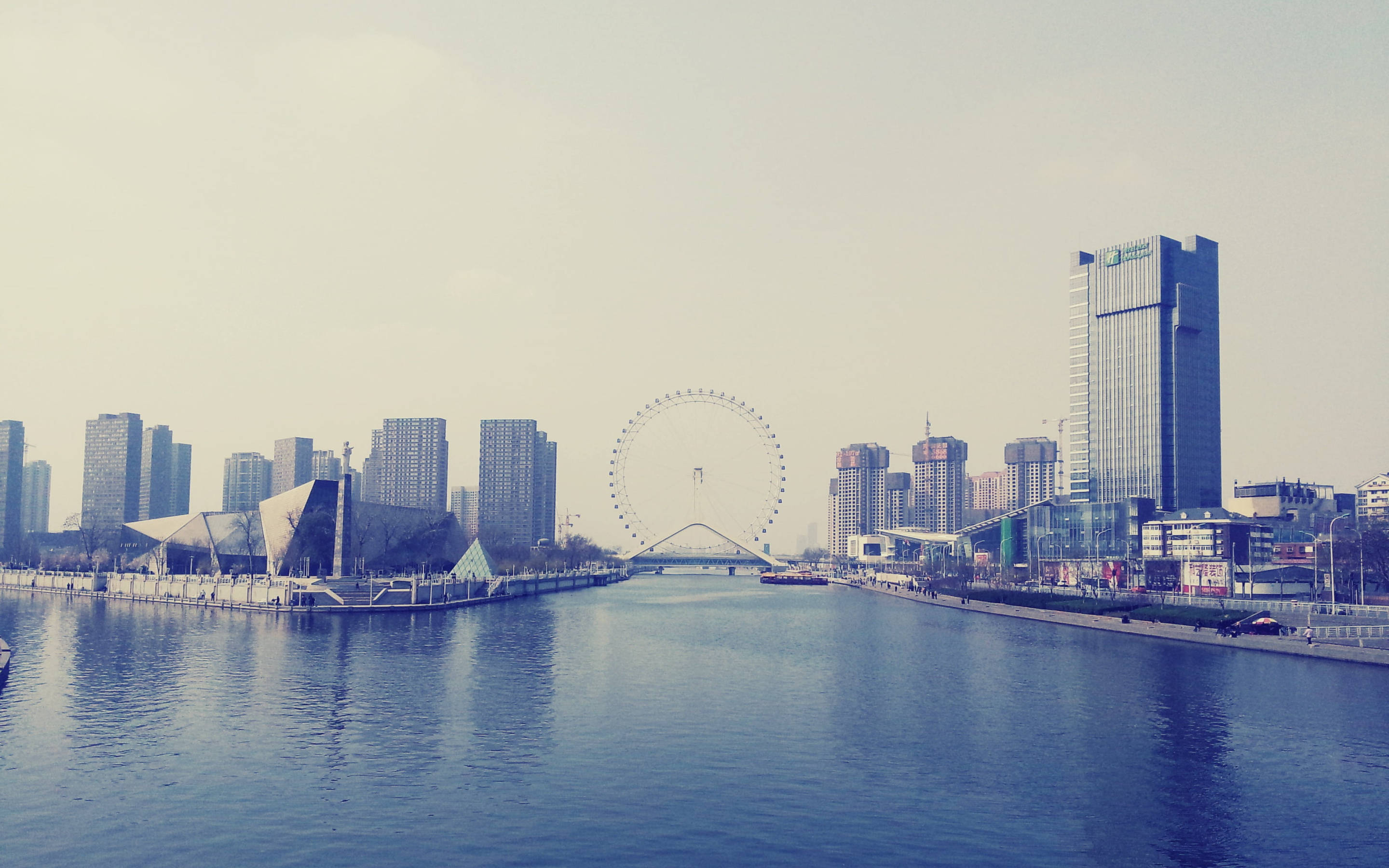 Tianjin 桌布 壁紙 — 一條平靜的河流流經現代城市景觀，高樓大廈和一個大型摩天輪在朦朧的天空下。