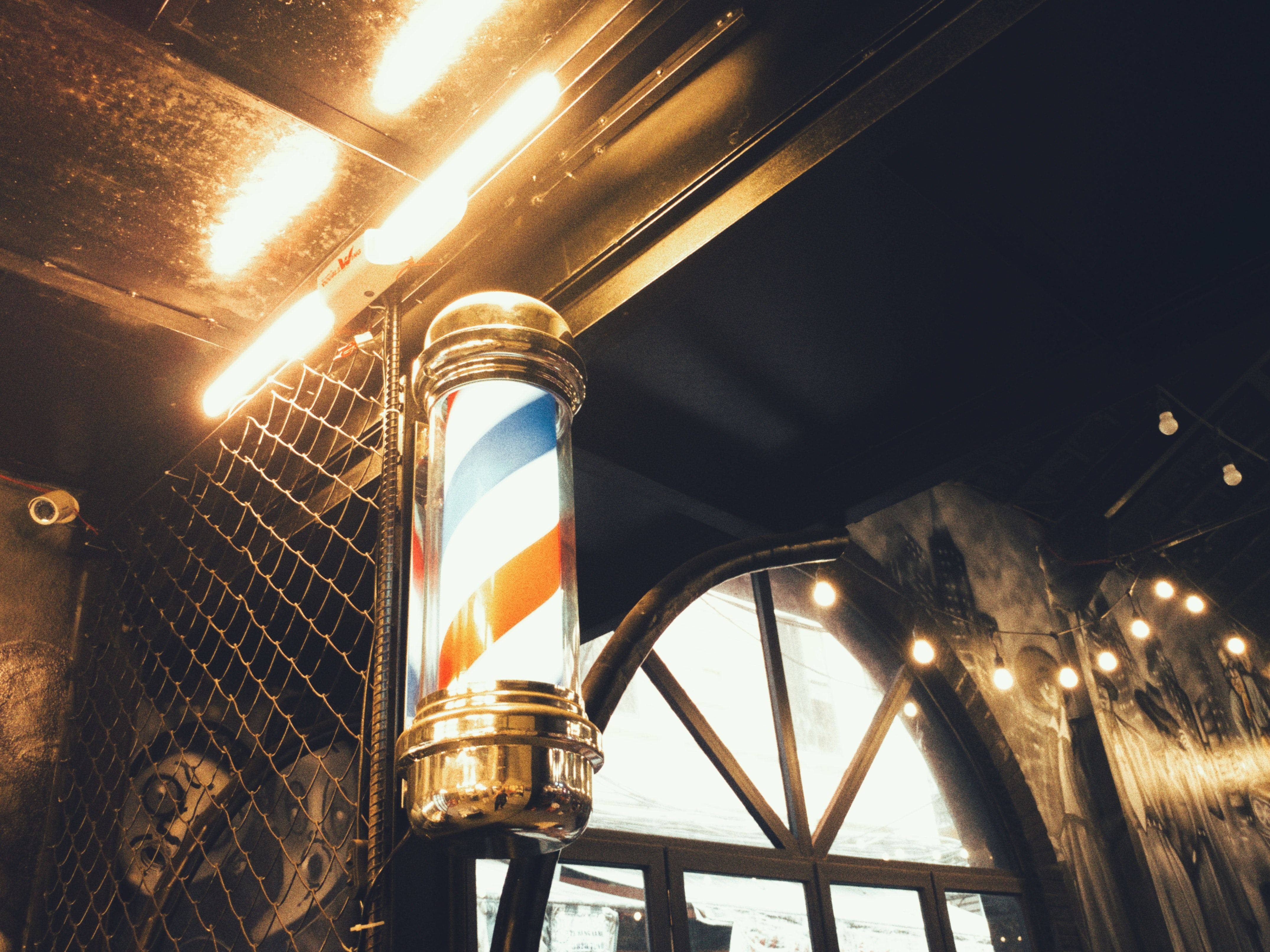 Barber 桌布 壁紙 — 一條復古理髮店旋轉燈在明亮的吊燈下溫暖地發光，位於一間有拱形窗戶和燈串的鄉村風格理髮店內。