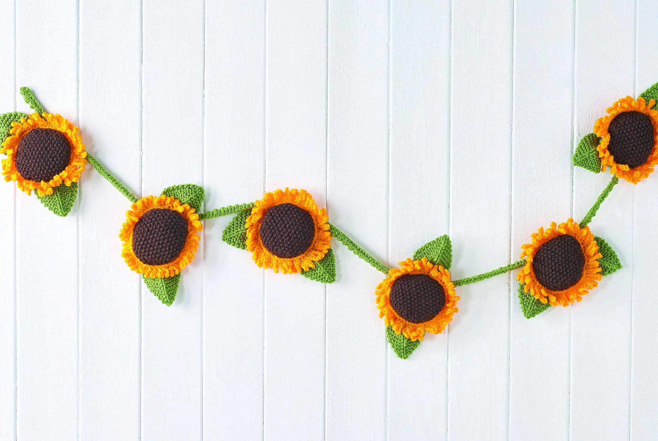 Sunflowers 桌布 壁紙 — 一條快樂的鉤針編織向日葵花環，帶有鮮豔的橙色花瓣和綠色葉子，掛在白色木質背景上。