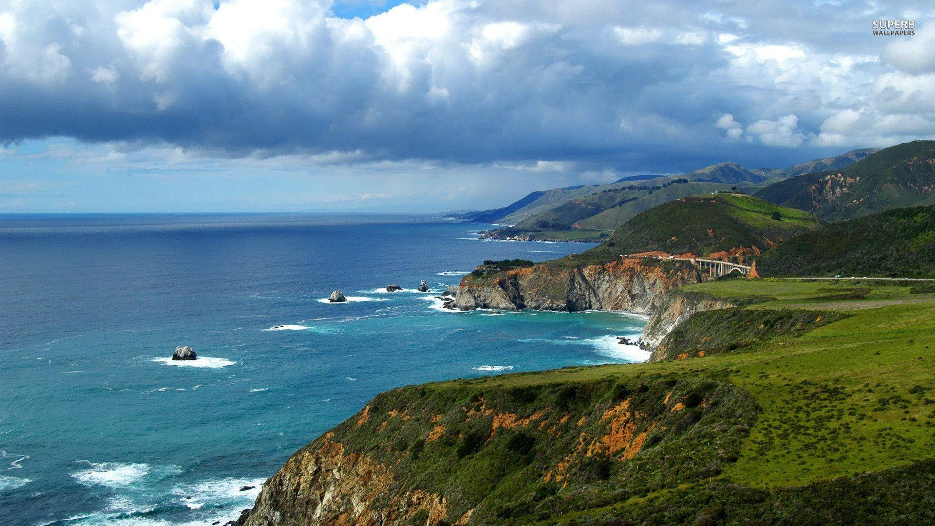 Big Sur 桌布 壁紙 — 一條戲劇性的海岸公路蜿蜒在崎嶇的懸崖邊，暴風雨的天空下，碧綠的海水拍打岩石。