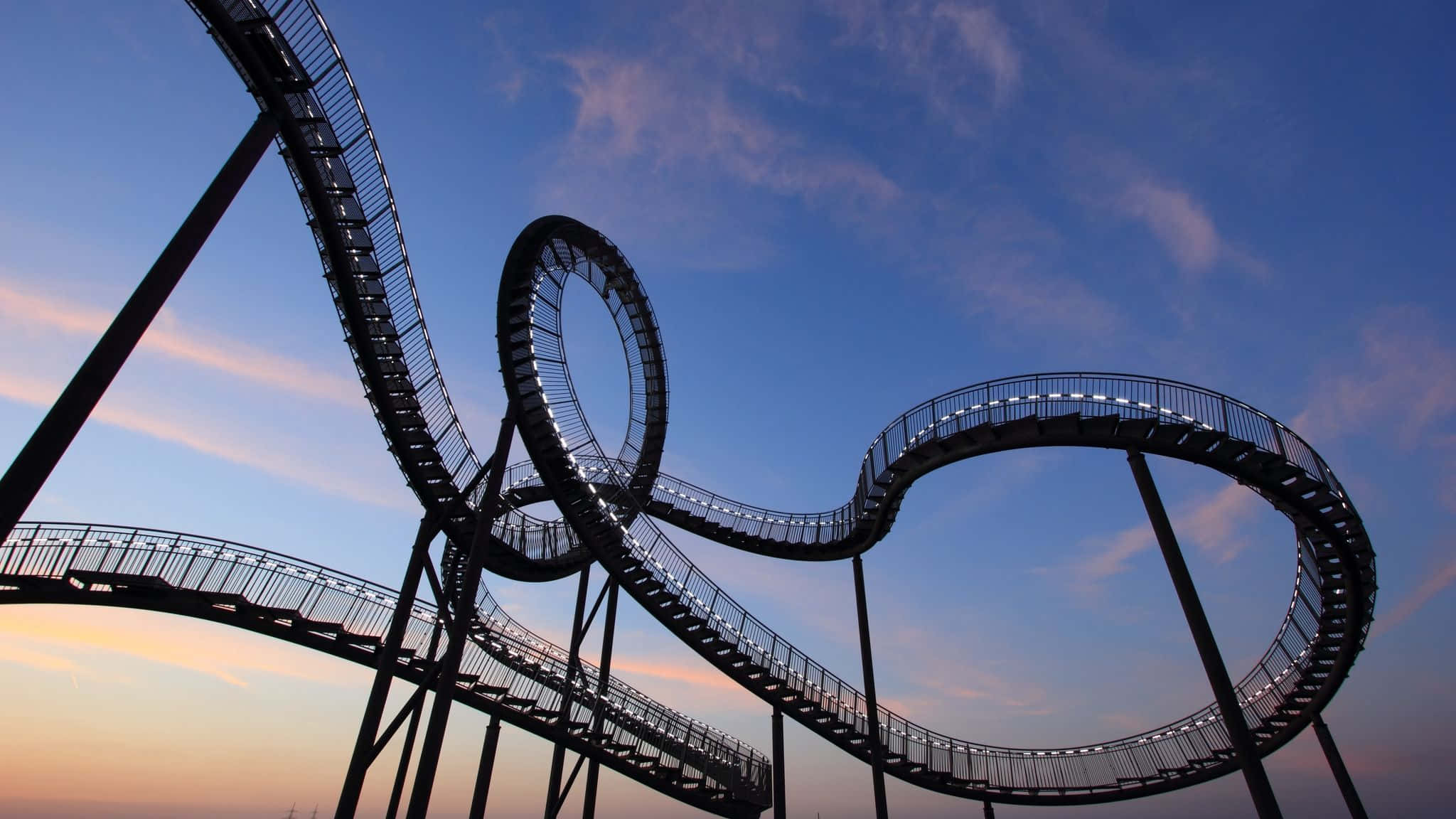 Roller Coaster 桌布 壁紙 — 一條扭曲的金屬空中步道，帶有發光的燈光，在帶有柔和粉紅色和藍色色調的暮色天空中彎曲。