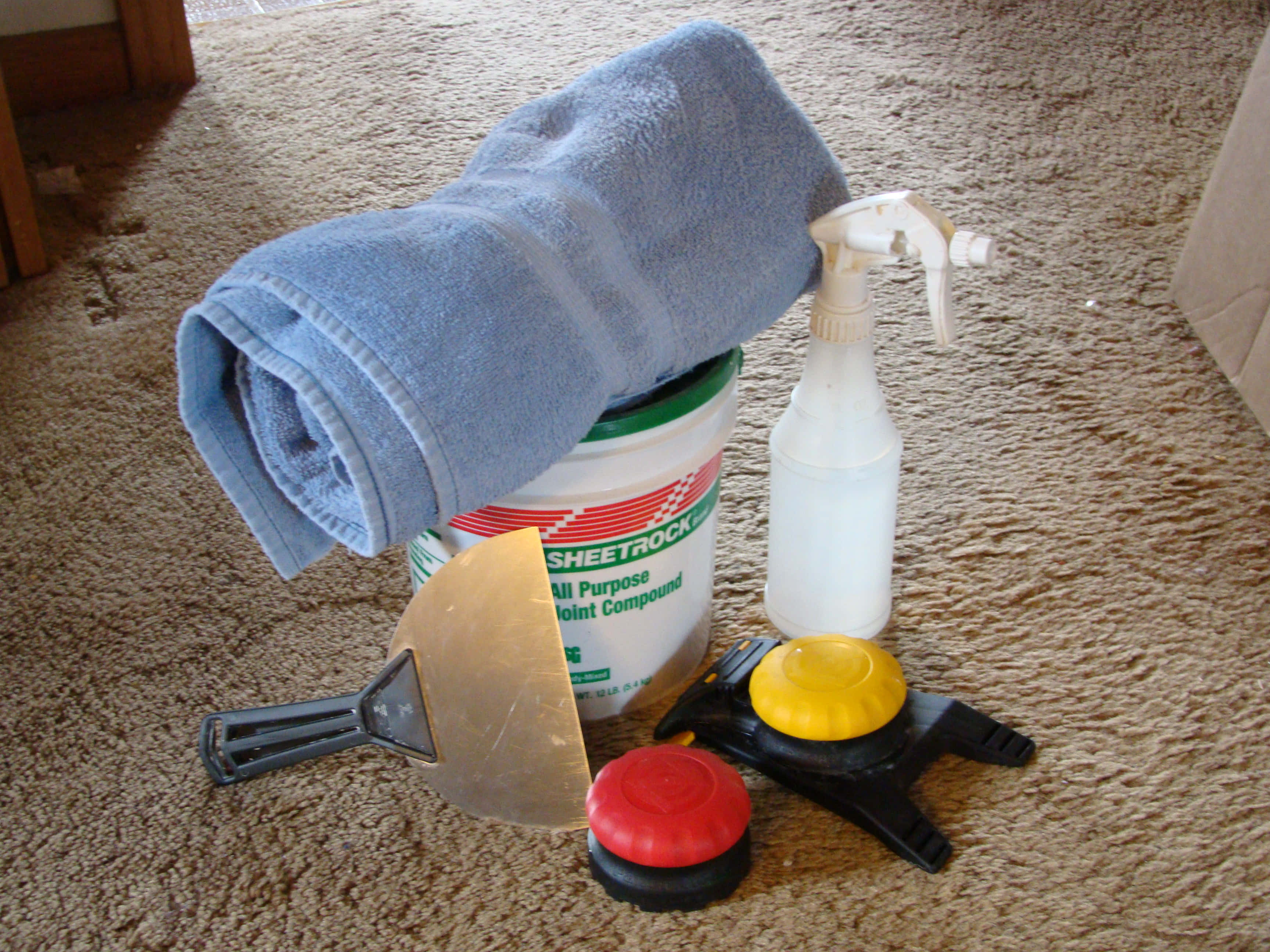 Cleaning 桌布 壁紙 — 一條捲起的藍色毛巾放在一桶接縫化合物上，旁邊有油灰刀、噴霧瓶和砂磨塊，位於米色地毯上。