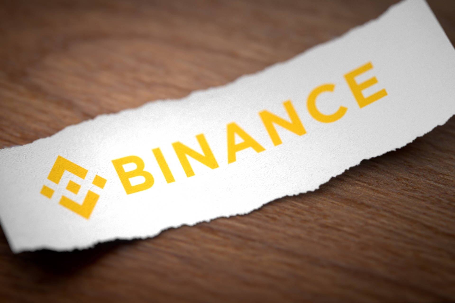 Binance 桌布 壁紙 — 一條撕裂的白色紙條帶有黃色 Binance 標誌，放在溫暖的木質表面上，景深較淺。