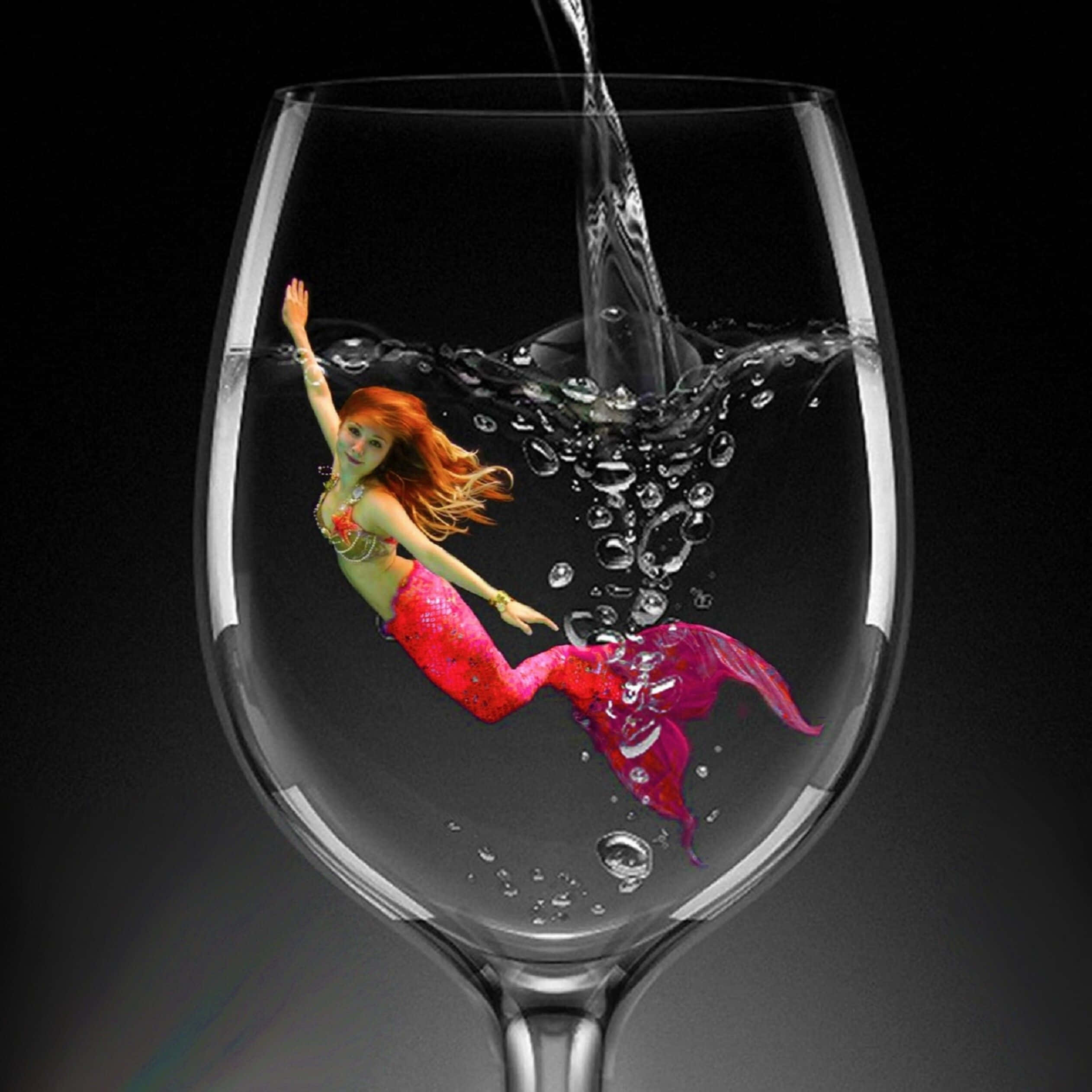 Glass Of Water 桌布 壁紙 — 一條擁有紅色尾巴的活潑美人魚在酒杯中優雅地游動，液體倒入時產生動態氣泡，背景為深色。