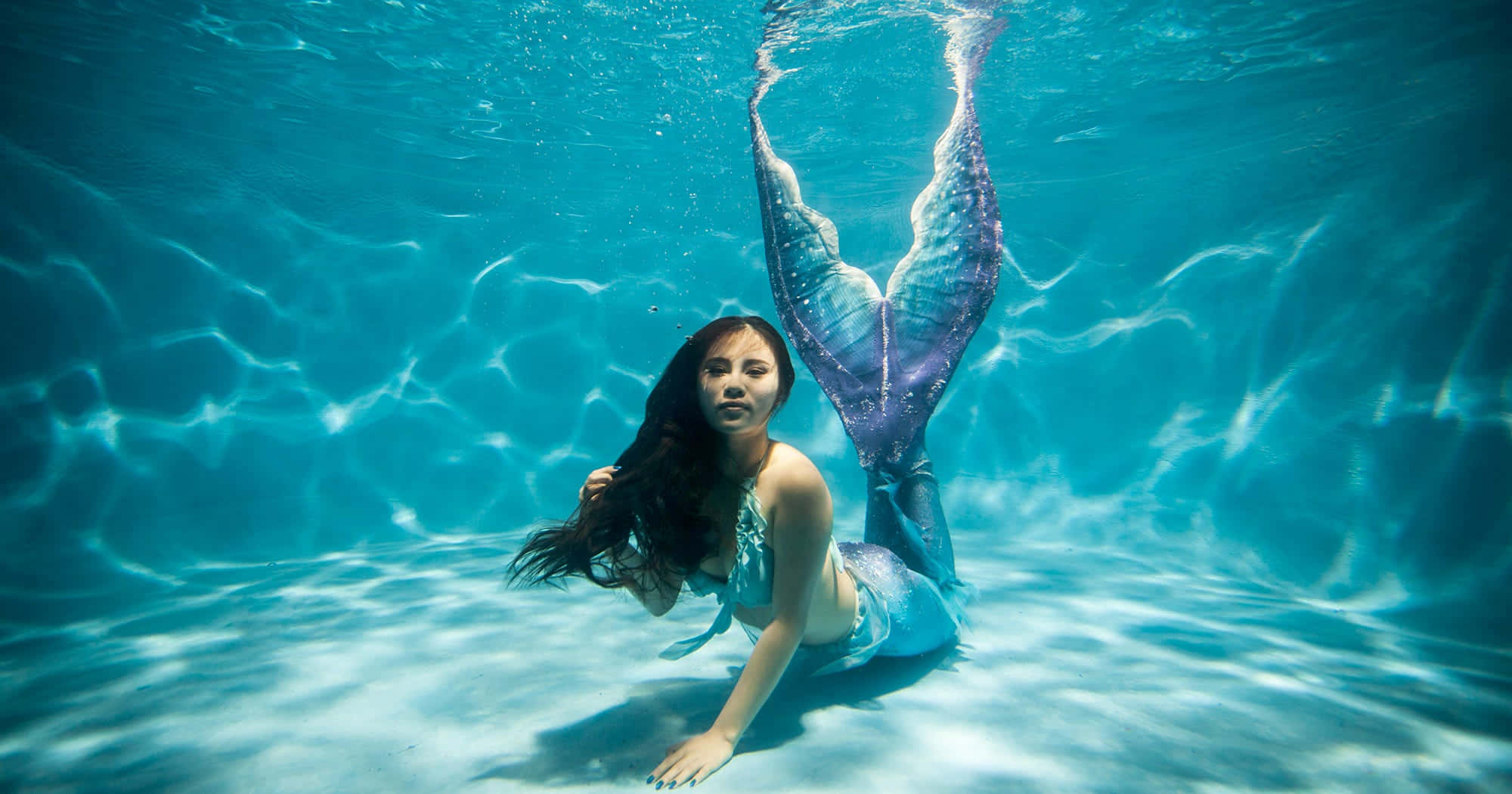 Real Mermaid 桌布 壁紙 — 一條擁有閃爍紫色尾巴的美人魚優雅地躺在泳池底部，陽光透過清澈的藍色水面照射進來。