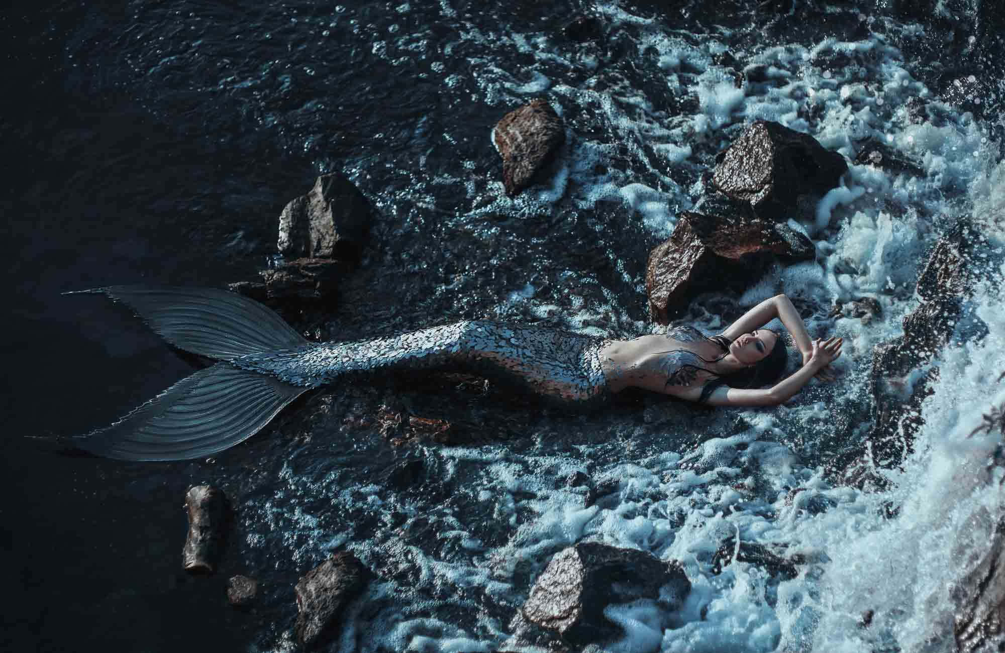 Mermaids 桌布 壁紙 — 一條擁有閃爍銀色尾巴的美人魚在波濤洶湧的海水中，於黑色岩石旁平靜地休息。