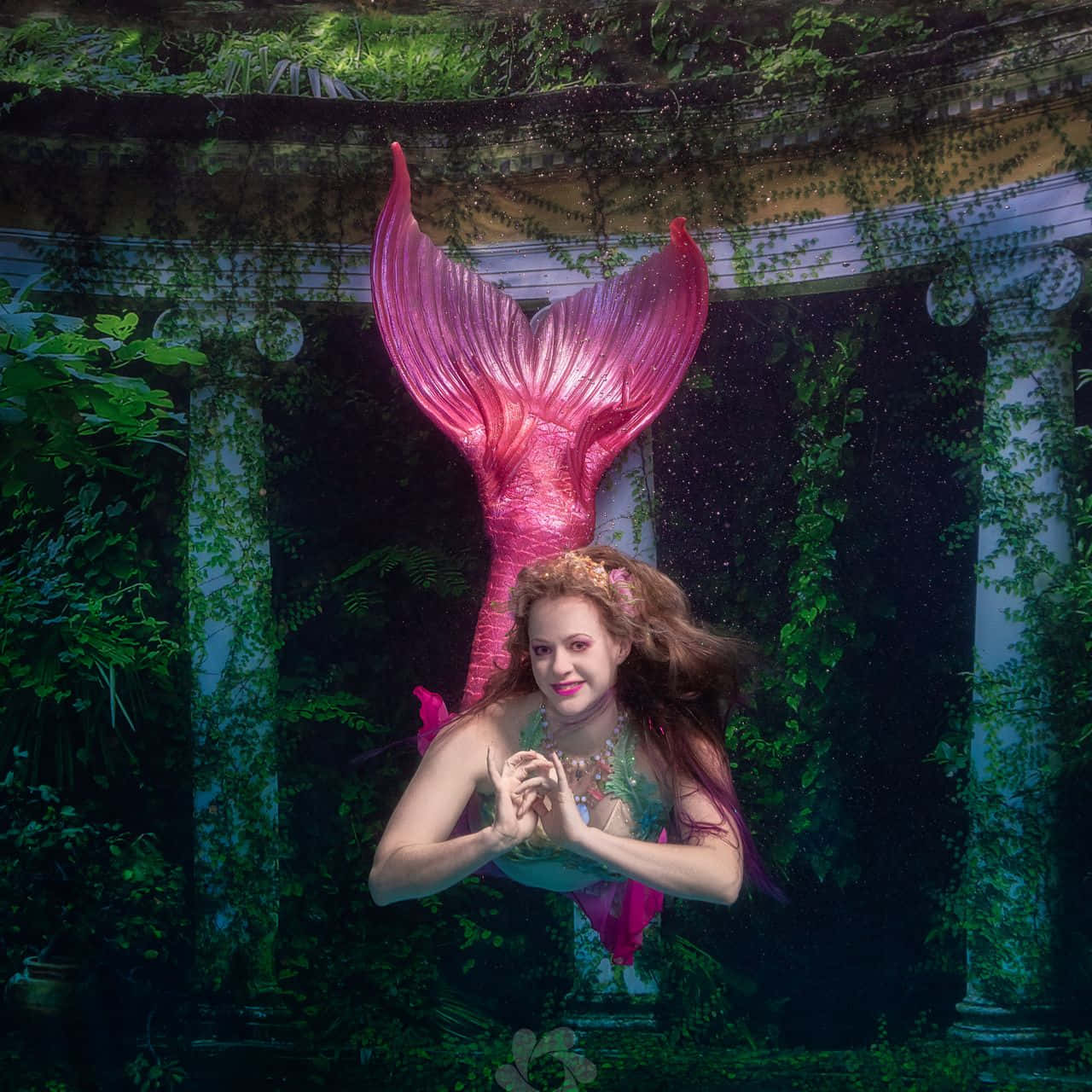 Mermaids 桌布 壁紙 — 一條擁有鮮豔粉紅色尾巴的美人魚從水中浮現，周圍環繞著茂密的綠植和古老的石柱。