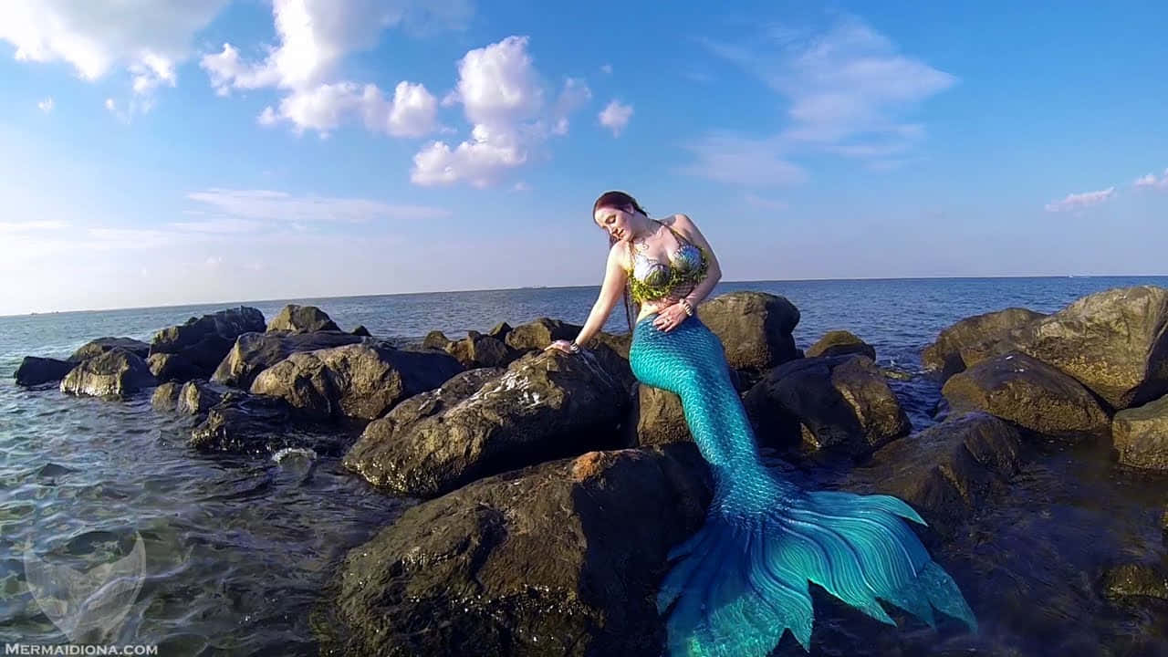 Mermaids 桌布 壁紙 — 一條擁有鮮豔藍色尾巴的美人魚，在明亮的藍天和蓬鬆白雲下，優雅地坐在海邊的深色岩石上。