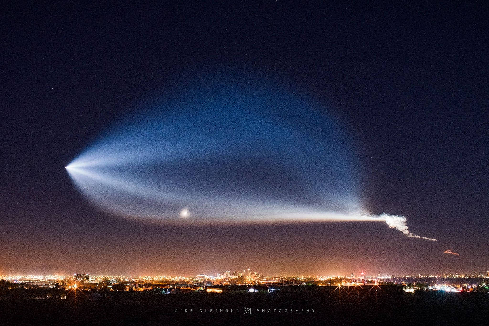 SpaceX 桌布 壁紙 — 一條明亮的藍色凝結尾跡劃過繁星點點的夜空，下方是發光的城市景觀。