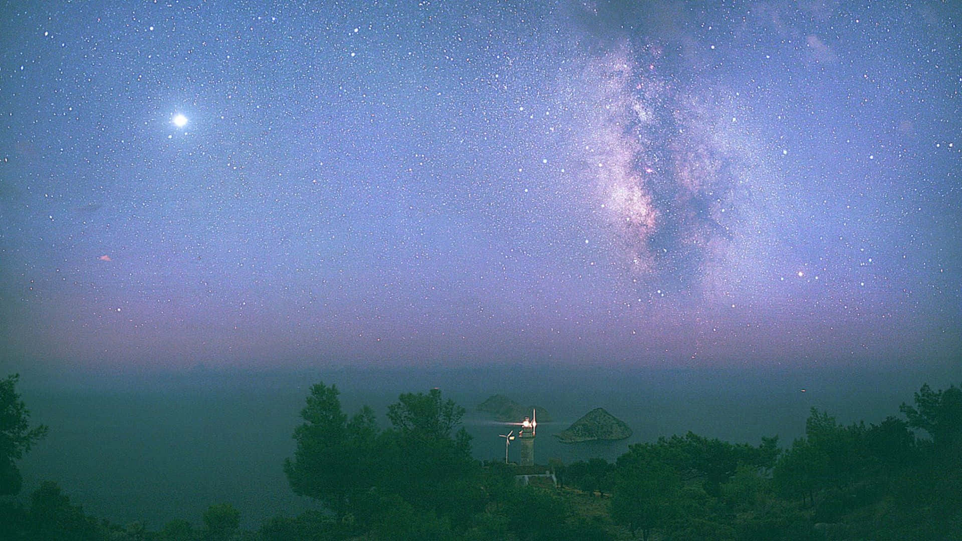 月亮與星星 桌布 壁紙 — 一條明亮的銀河在霧氣瀰漫的海岸景觀上空拱起，有一座發光的燈塔和剪影樹木，背景是星光閃爍的黃昏天空。