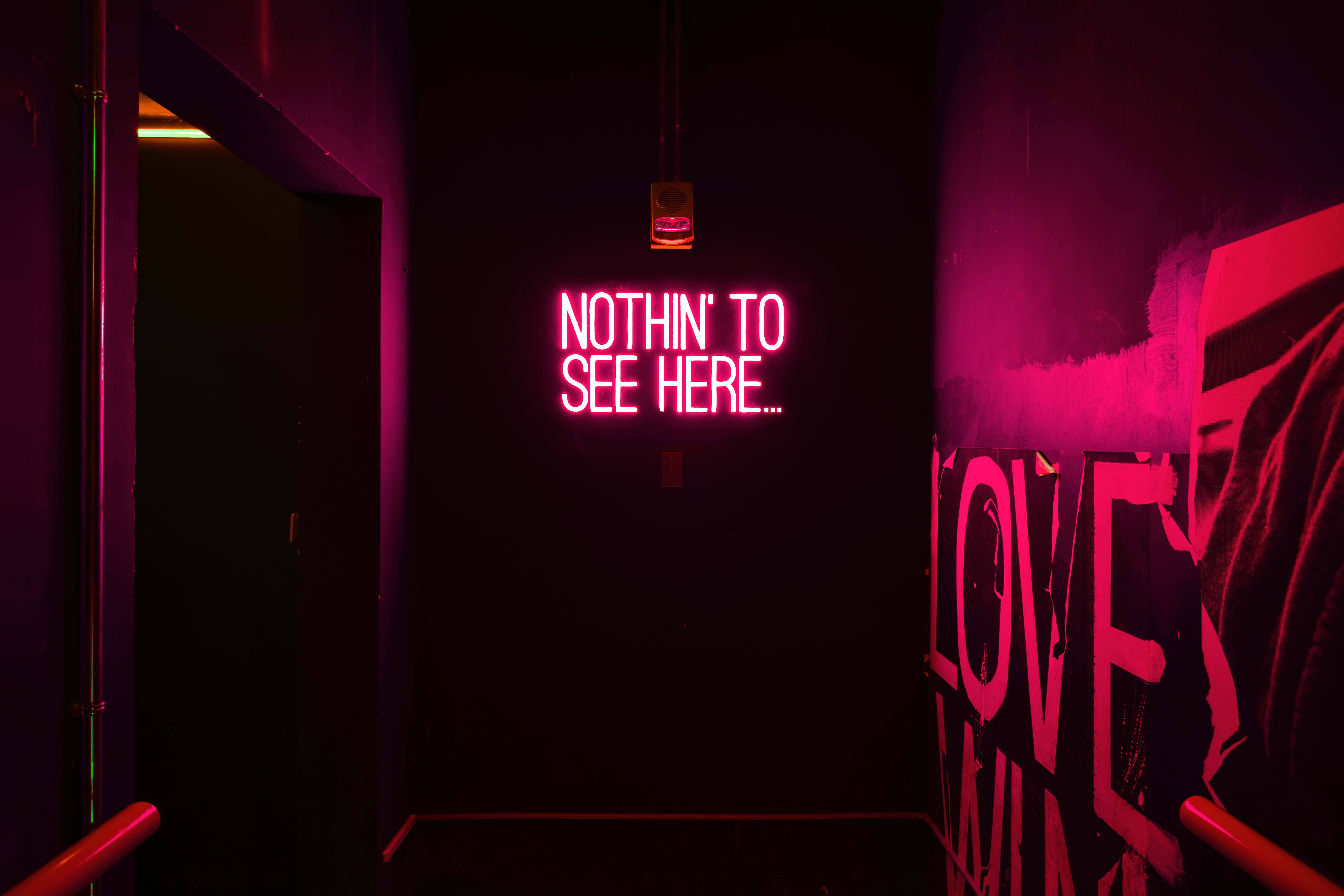 Neon Red Aesthetic 桌布 壁紙 — 一條昏暗的走廊沐浴在洋紅色燈光中，門口上方有一個發光的霓虹燈牌，上面寫著「NOTHIN' TO SEE HERE...」。