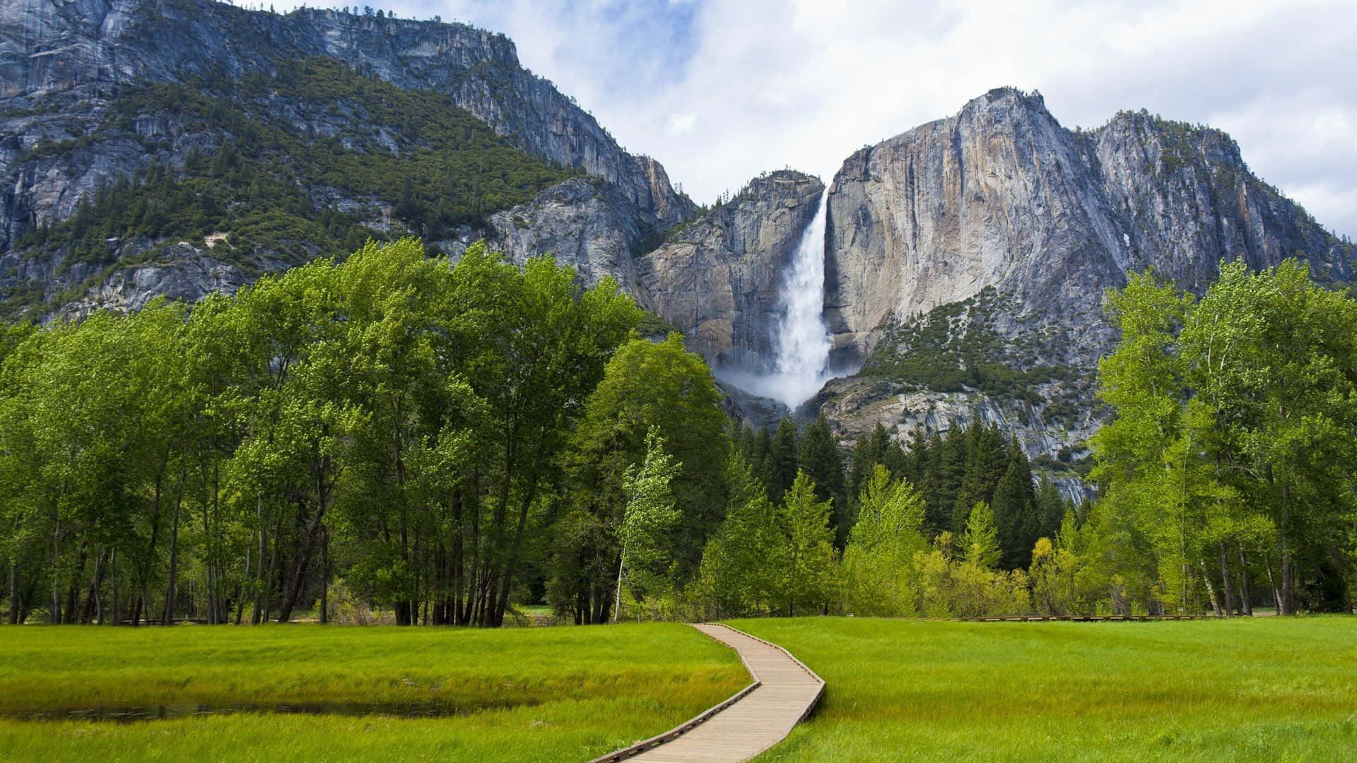 Waterfalls 桌布 壁紙 — 一條木棧道蜿蜒穿過充滿活力的綠色草地，通往從崎嶇花崗岩懸崖傾瀉而下的雄偉瀑布。