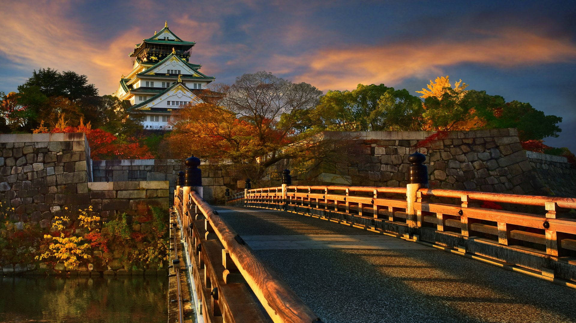Osaka Castle 桌布 壁紙 — 一條木橋通往日落時分雄偉的日本城堡，周圍有鮮豔的秋葉和溫暖的金色光芒。