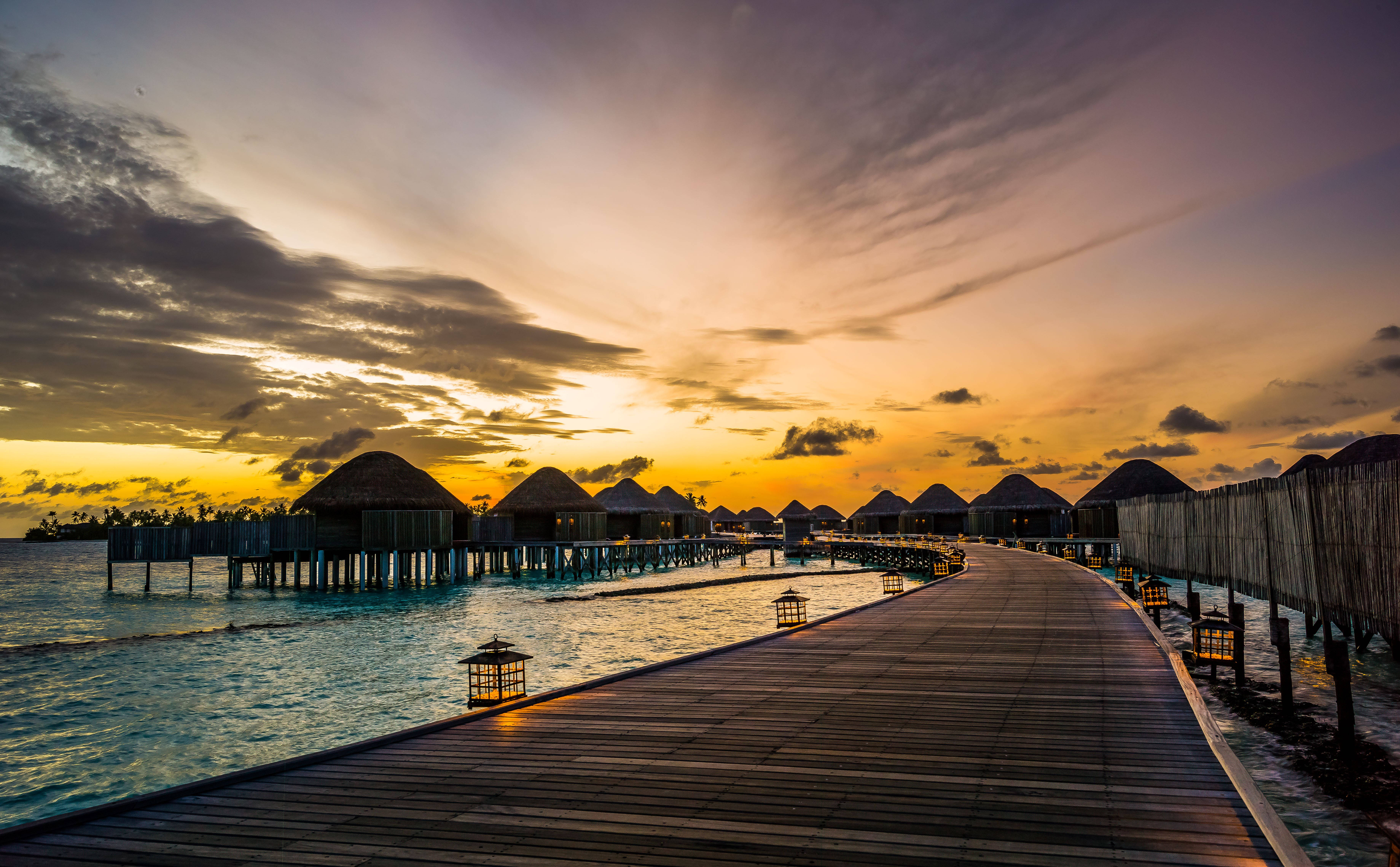 Maldives 桌布 壁紙 — 一條木製棧道在日落時分延伸向水上平房，金色天空與平靜的碧綠海水相映。