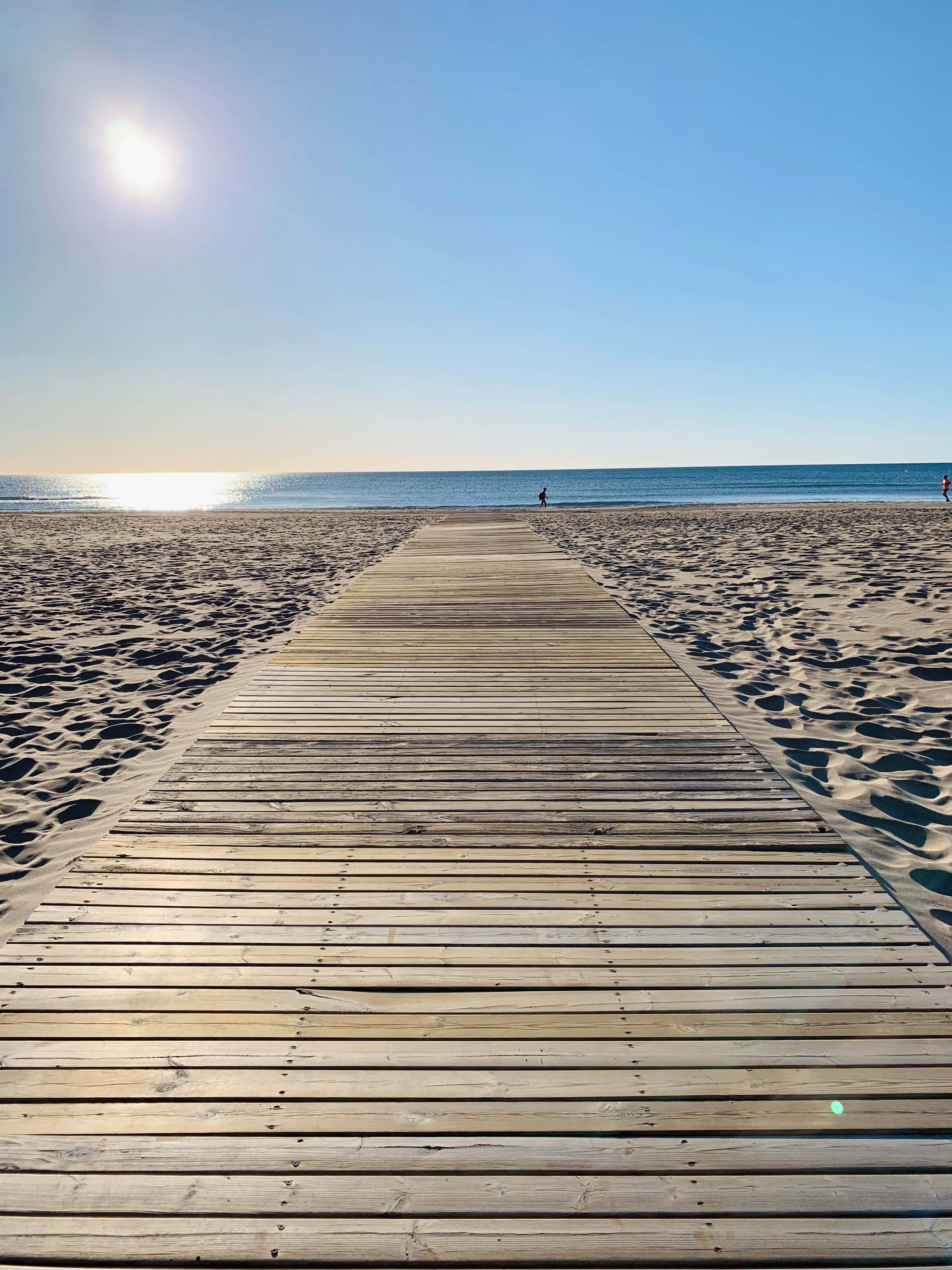Boardwalk 桌布 壁紙 — 一條木製棧道在明媚陽光下延伸至海洋，天空湛藍，遠處有海灘遊客。