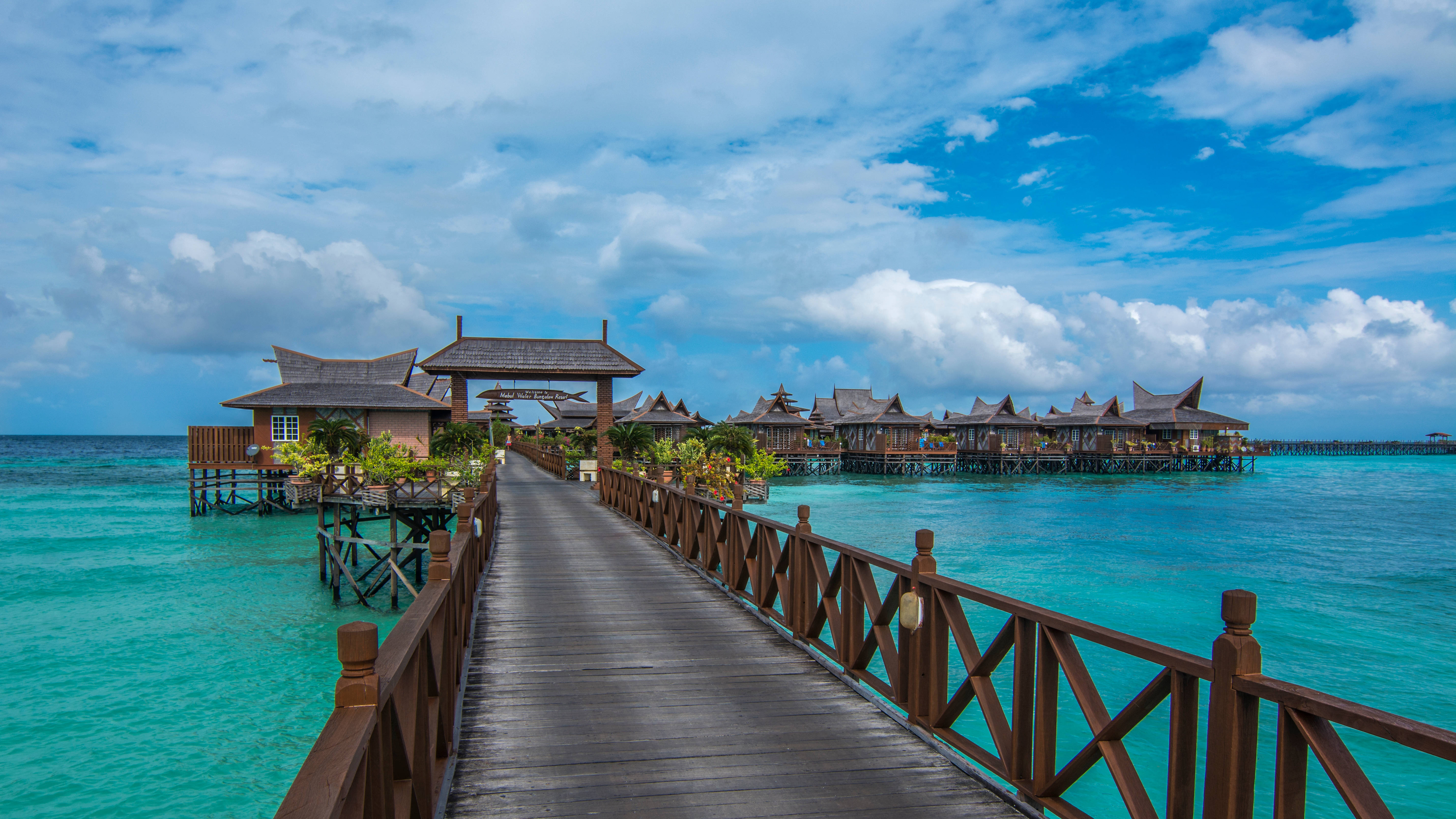 Malaysia 桌布 壁紙 — 一條木製棧道延伸在碧綠色的海水上，通往傳統的水上平房，上方是晴朗的藍天和白雲。