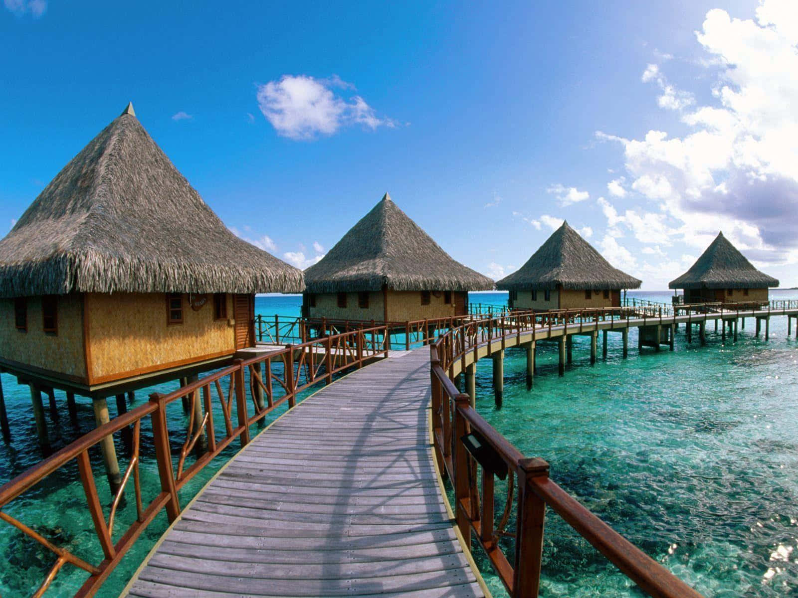 Bora Bora 桌布 壁紙 — 一條木製棧道蜿蜒穿過碧綠海水，通往茅草屋頂的水上平房，上方是藍天白雲。