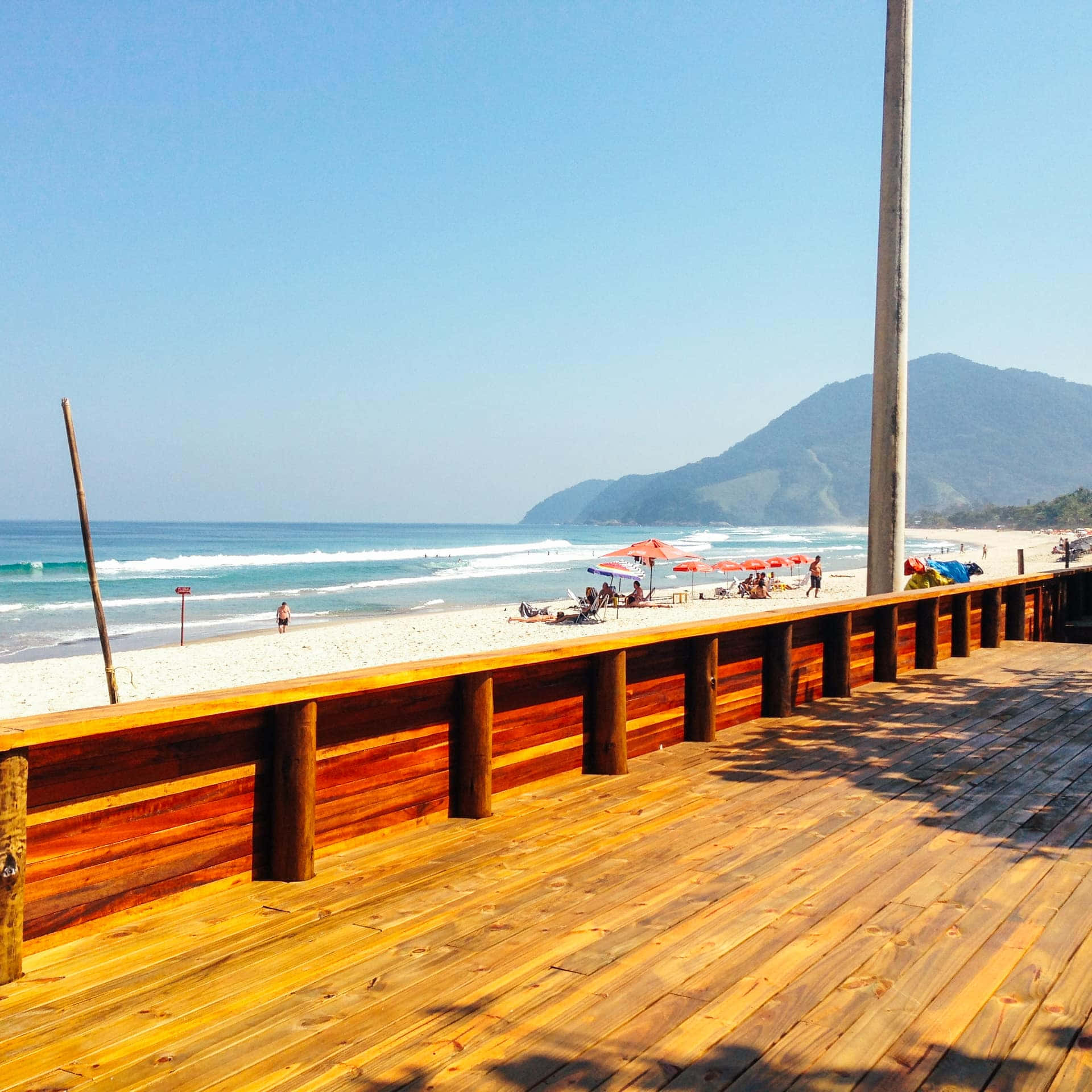 Brazilian Beach 桌布 壁紙 — 一條木製步道俯瞰著陽光明媚的海灘，碧綠色的海浪拍打著白色沙灘和遠處的綠色山丘，天空清澈湛藍。