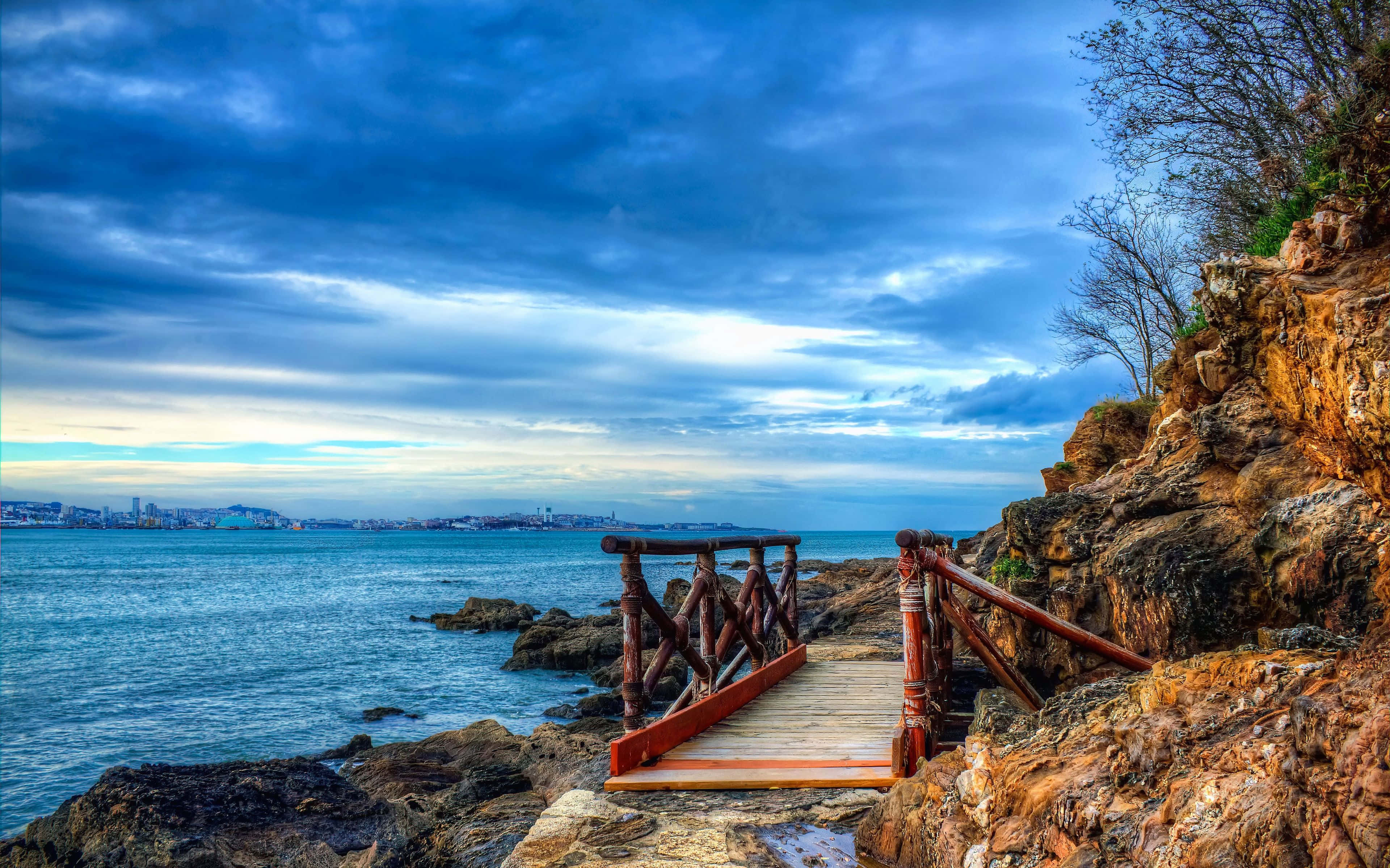 4K Hdr 桌布 壁紙 — 一條木製步道沿著崎嶇的懸崖蜿蜒而行，俯瞰廣闊的藍色大海，上方是戲劇性的多雲天空。