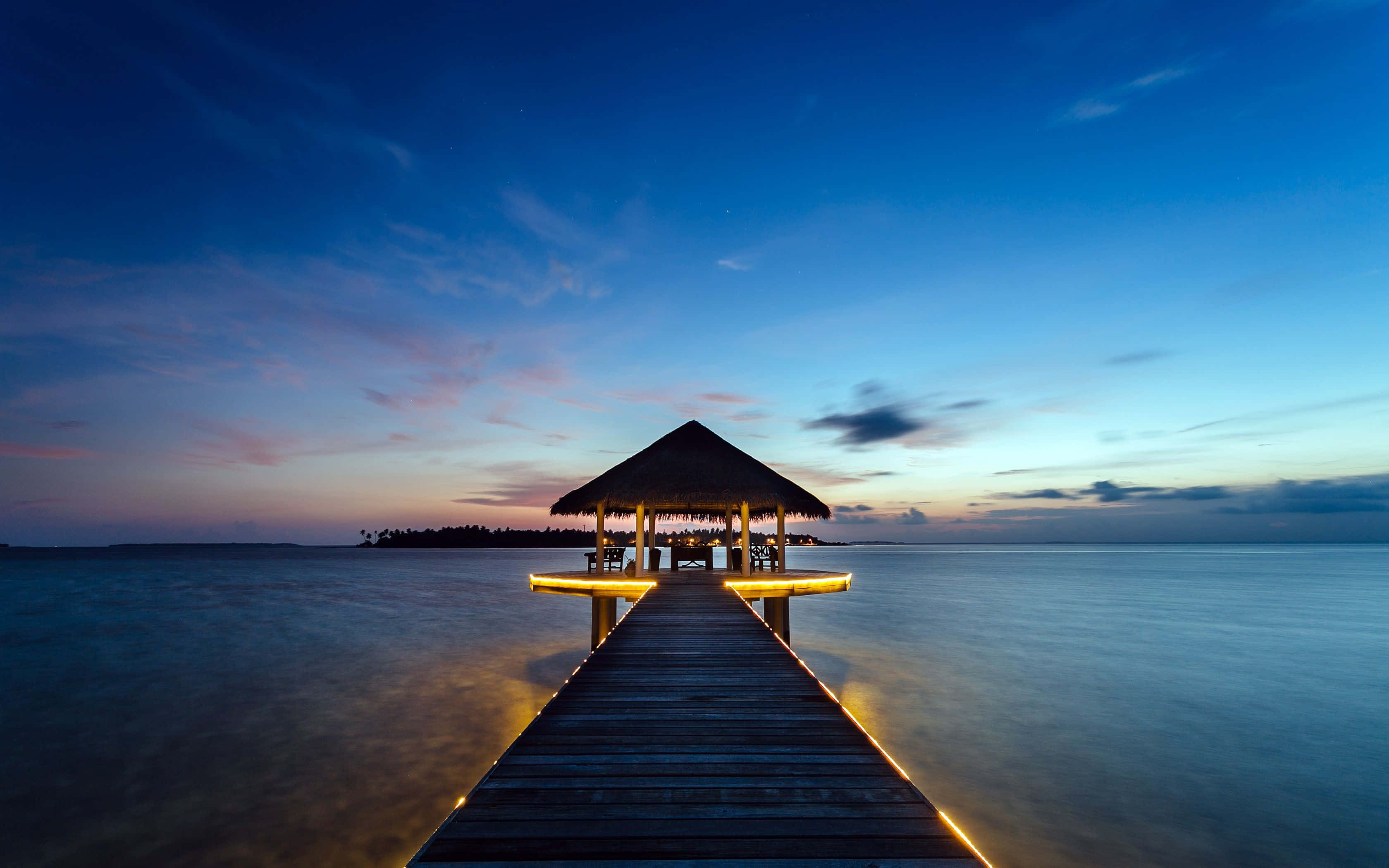 Maldives Beach 桌布 壁紙 — 一條木製碼頭延伸至平靜的海水，通往一個在黃昏時分散發溫暖光芒的茅草屋頂涼亭。