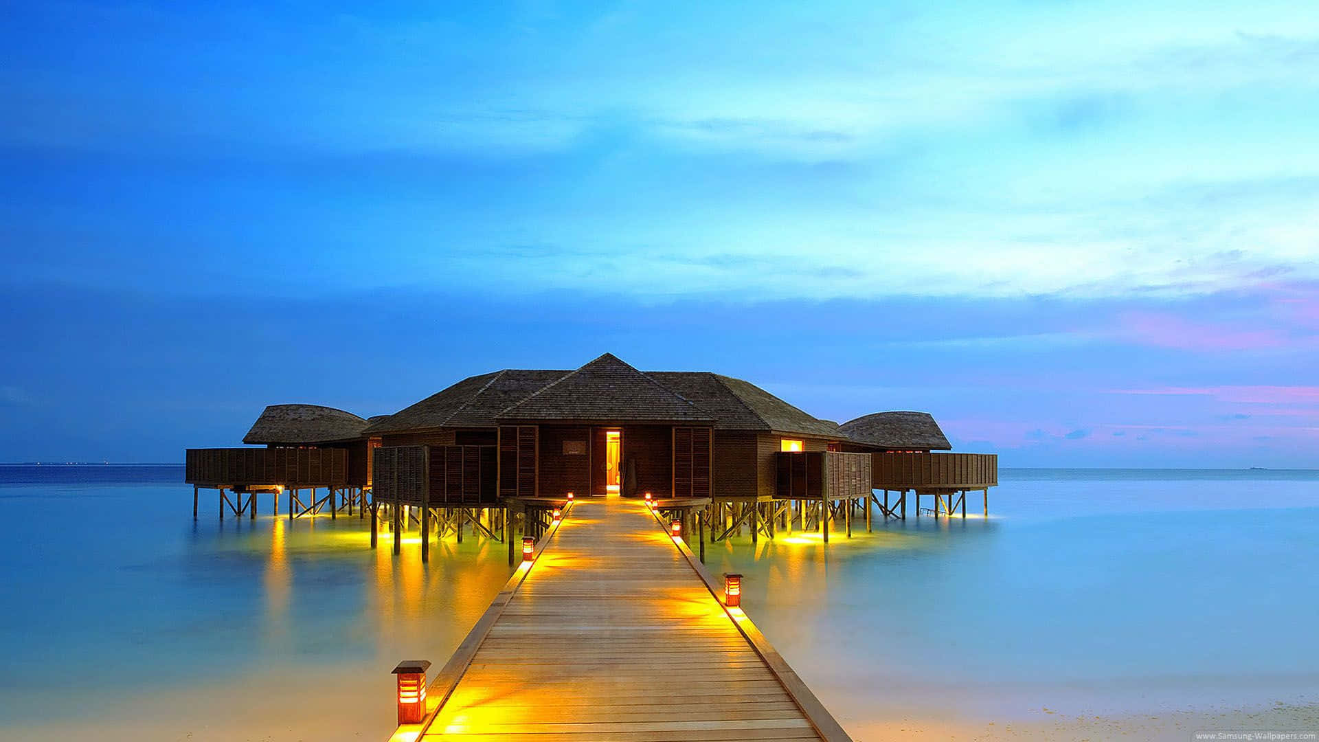 Maldives Beach 桌布 壁紙 — 一條木製碼頭通往黃昏時發光的水上平房，平靜的碧綠海水和柔和的藍天。