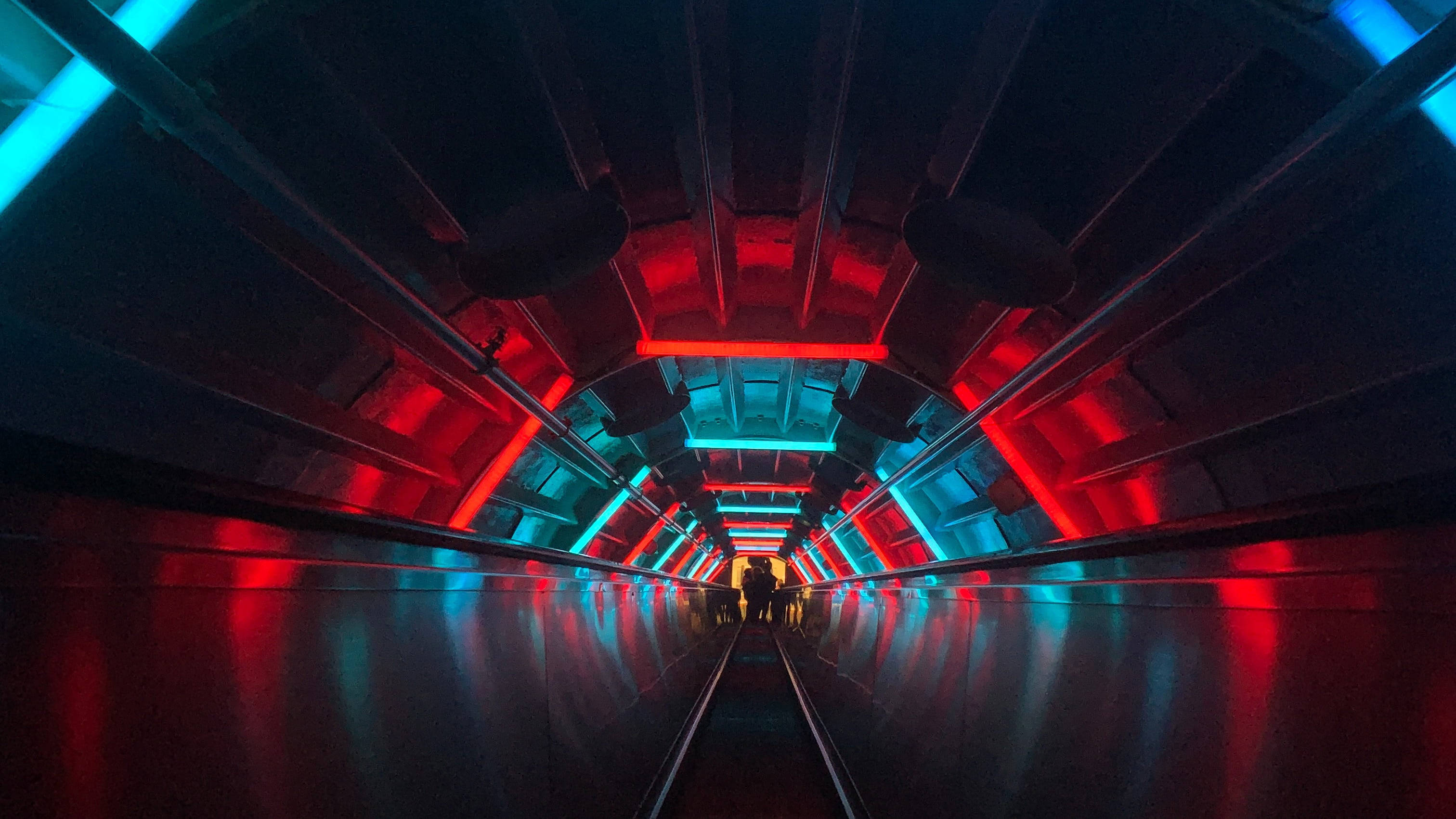 Dark Neon 桌布 壁紙 — 一條未來風格的隧道，紅色和藍色霓虹燈光在光澤地板上反射，剪影人物走向消失點。