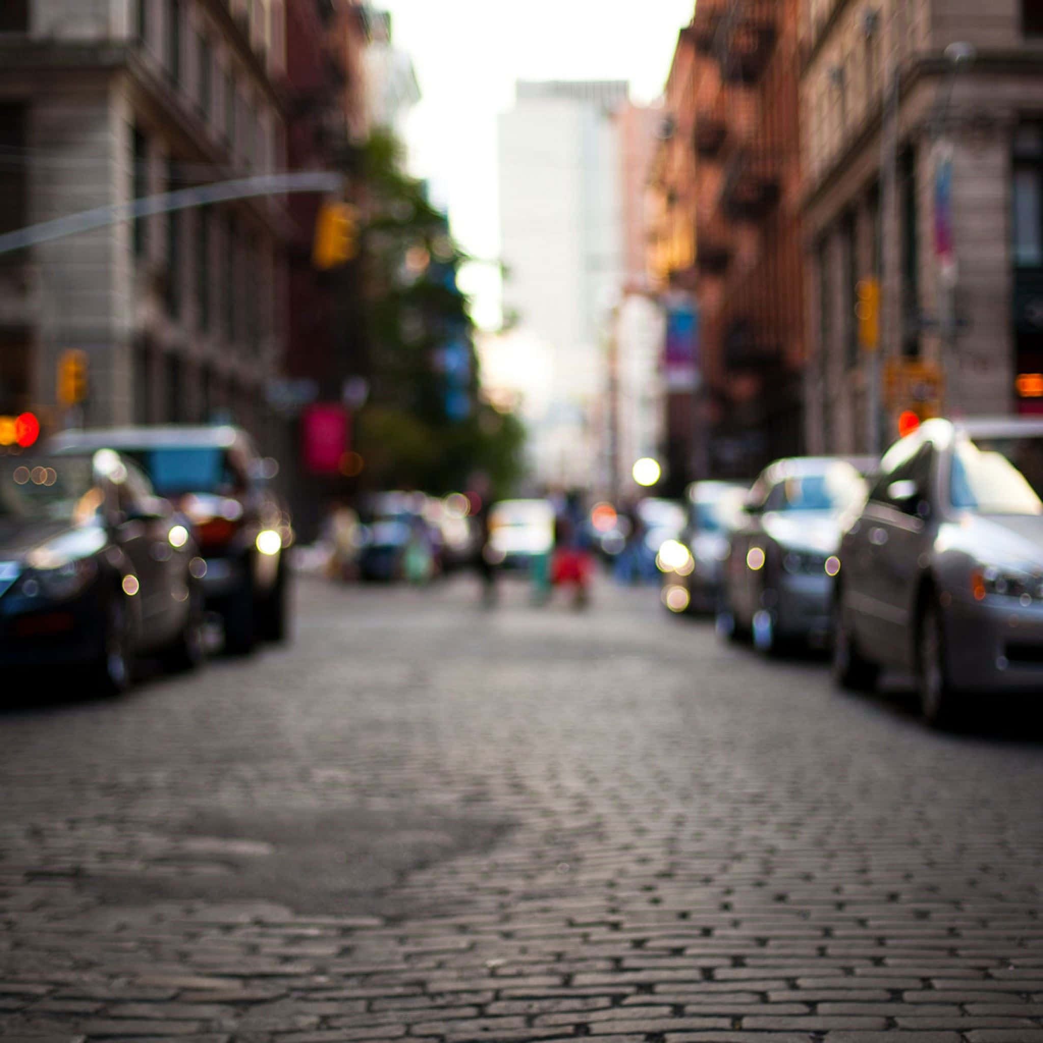 City Street 桌布 壁紙 — 一條模糊的鵝卵石街道，城市中有停泊的汽車和遠處的行人，在柔和日光下。