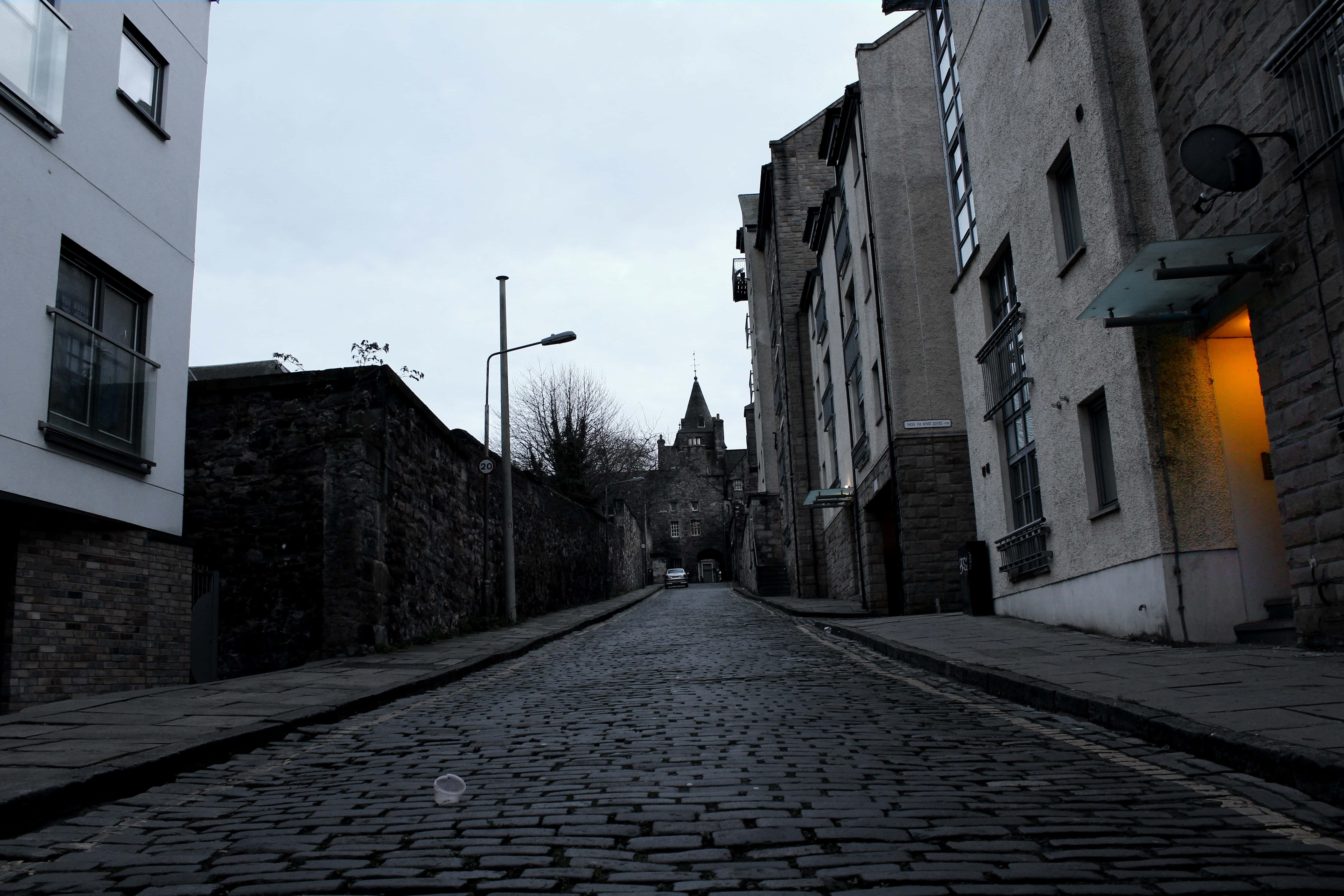Dark Alleyway 桌布 壁紙 — 一條歷史名城的狹窄鵝卵石街道，在陰天之下通往遠處的城堡，右邊有一道溫暖亮著的門口。