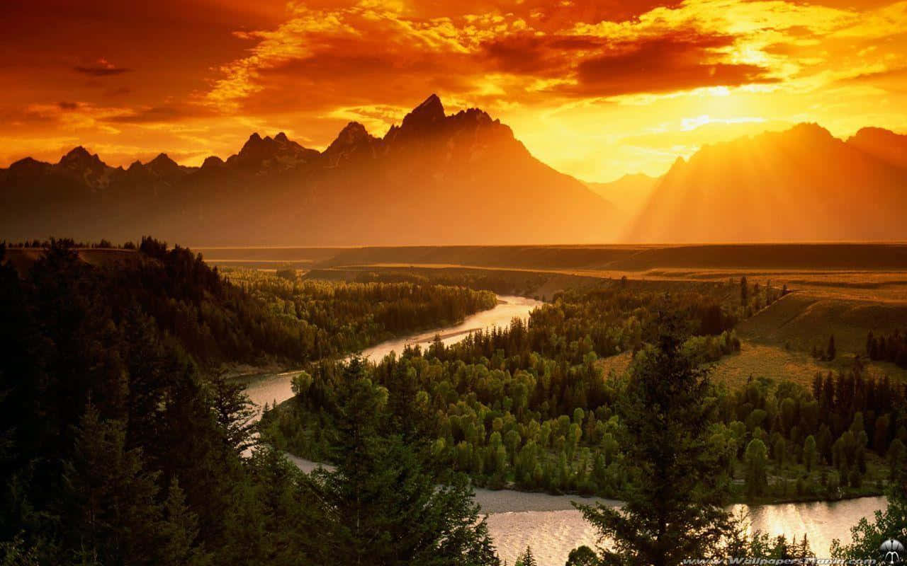 Yellowstone 桌布 壁紙 — 一條河流蜿蜒穿過森林覆蓋的山谷，金色陽光穿透雲層，照射在日落時分崎嶇的山峰之上。