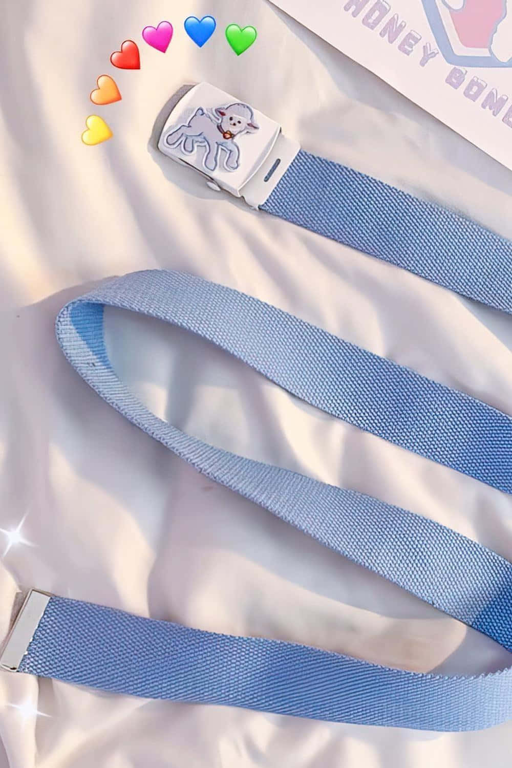 Pastel Blue 桌布 壁紙 — 一條淺藍色編織皮帶，帶有白色夾子，上面有羊的設計，放在柔軟的白色布料上，周圍環繞著彩色心形表情符號和一個「HONEY BOMB」標籤。